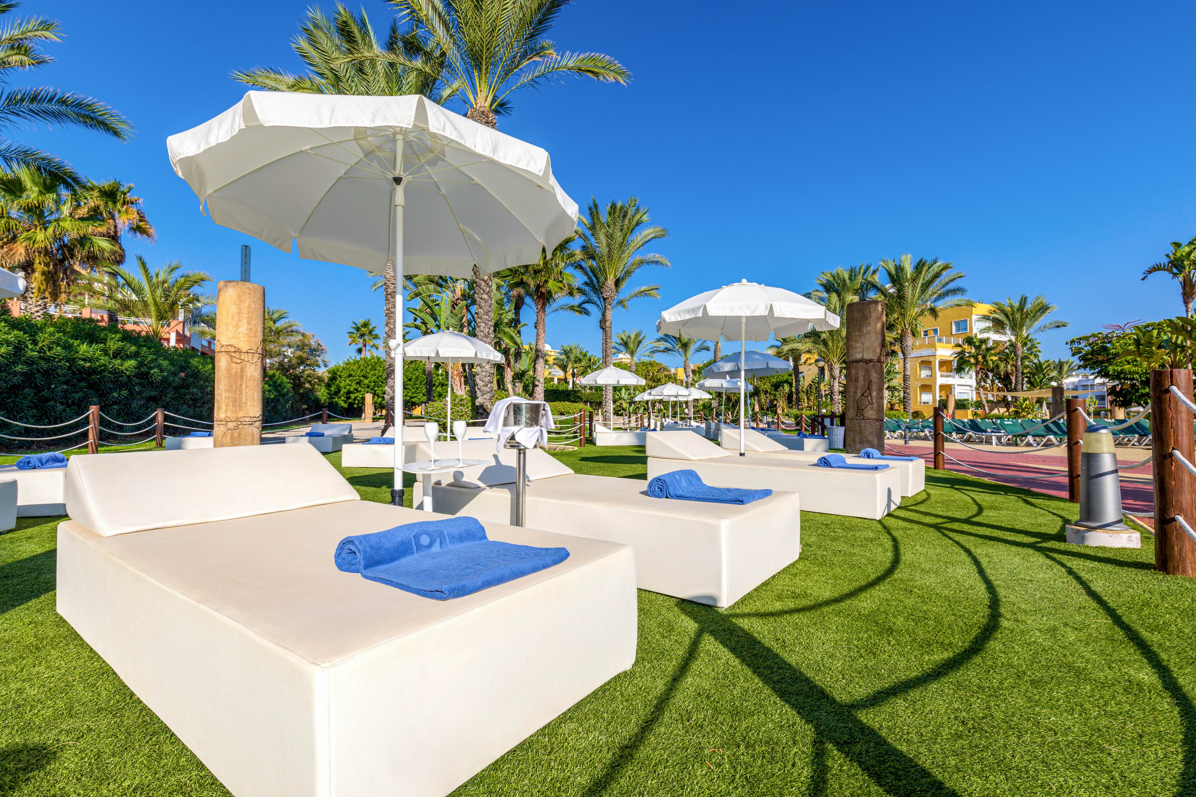 Outdoor resort lounge area with large white sunbeds and blue towels arranged on artificial grass. White parasol umbrellas provide shade under a clear blue sky. Palm trees and lush greenery surround the seating area, creating a tropical vacation atmosphere. In the background, more loungers and a poolside area suggest a relaxing holiday setting.
