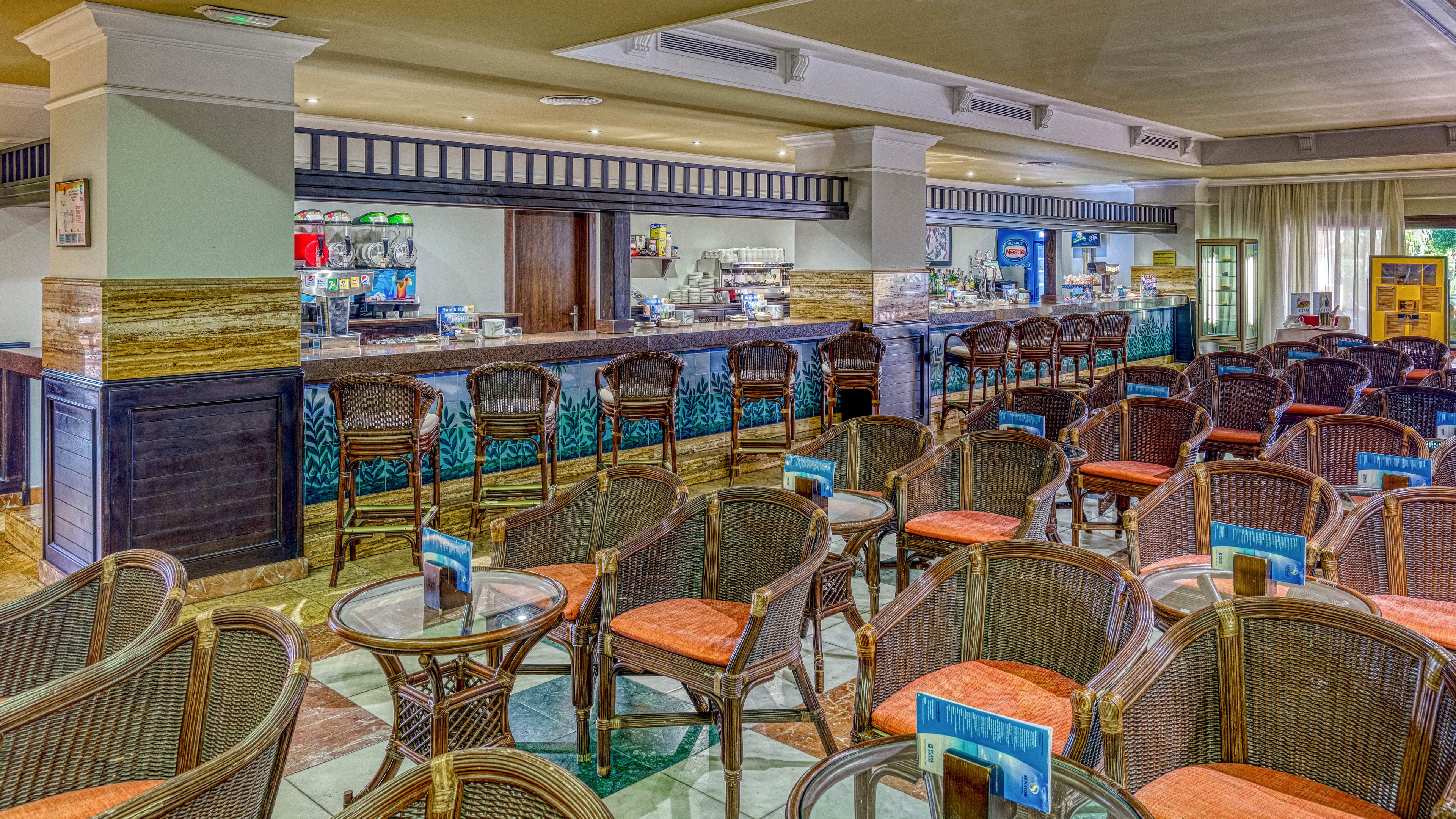 A spacious resort bar lounge features rows of wicker chairs with orange cushions arranged around small round tables. The long bar counter in the background is stocked with bottles, drink dispensers, and glassware. Bright interior lighting, tiled floors, and decorative wall details create a clean, inviting atmosphere. No people are present, emphasizing the orderly layout and relaxed vacation setting.