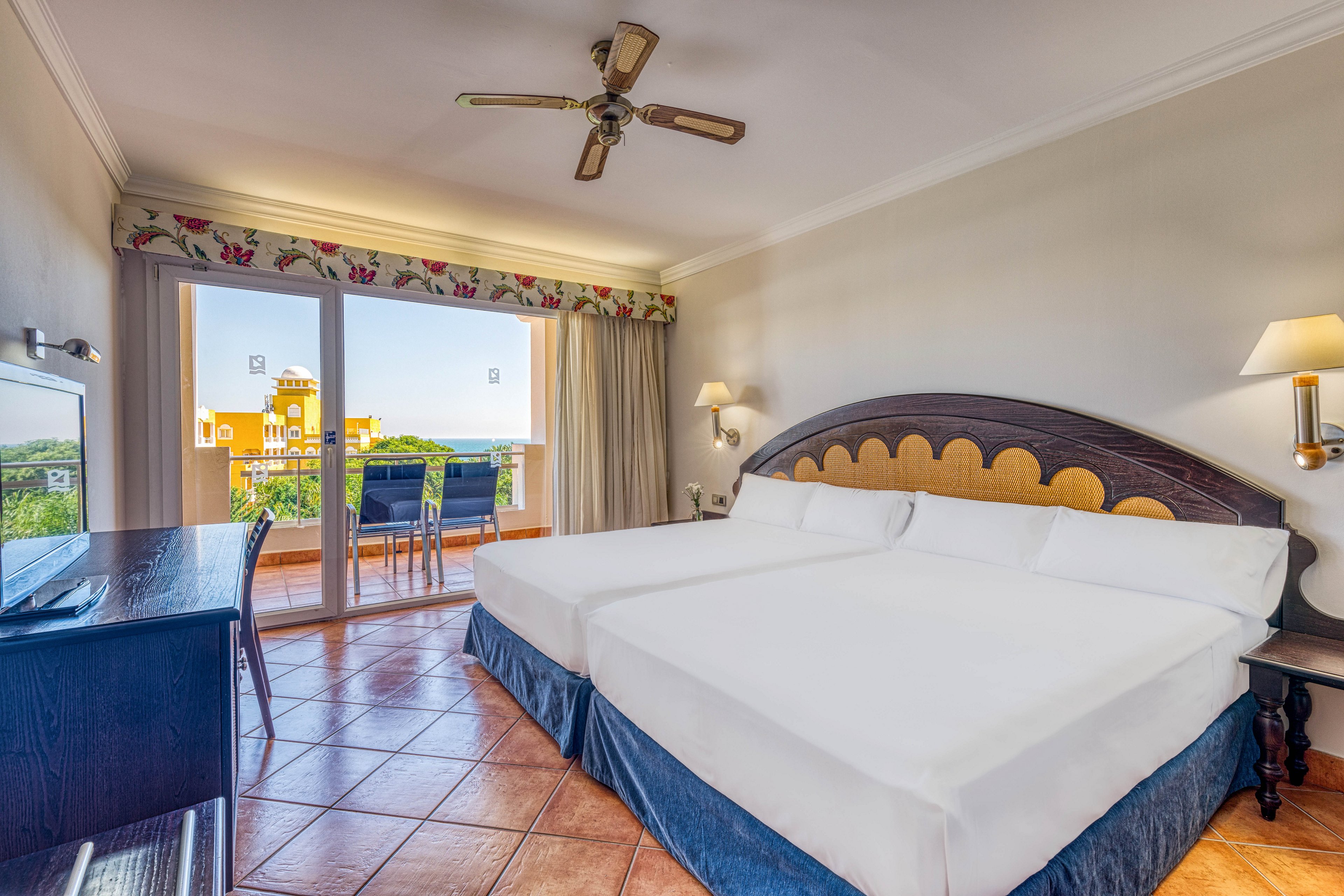 Spacious hotel room featuring two large beds with white linens and a dark wooden headboard. The room opens onto a terrace with a table and chairs overlooking colorful buildings and greenery. Warm terracotta floor tiles and neutral walls create a cozy, inviting atmosphere. A ceiling fan, wall lamps, and a desk with mirror complete the comfortable guestroom setting.