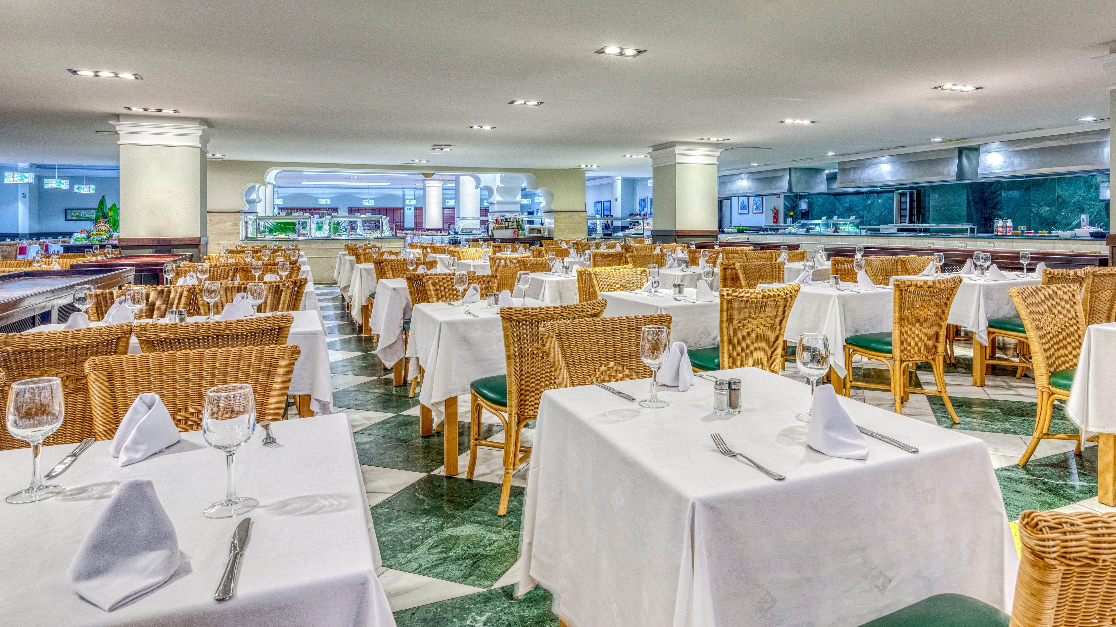 A spacious restaurant dining room is shown with rows of neatly arranged tables covered in white tablecloths. Each table is set with folded napkins, glassware, and cutlery, ready for guests. Wicker chairs with green cushions add warmth to the bright interior. The space is well lit with ceiling lights and features a long buffet or service counter in the background.