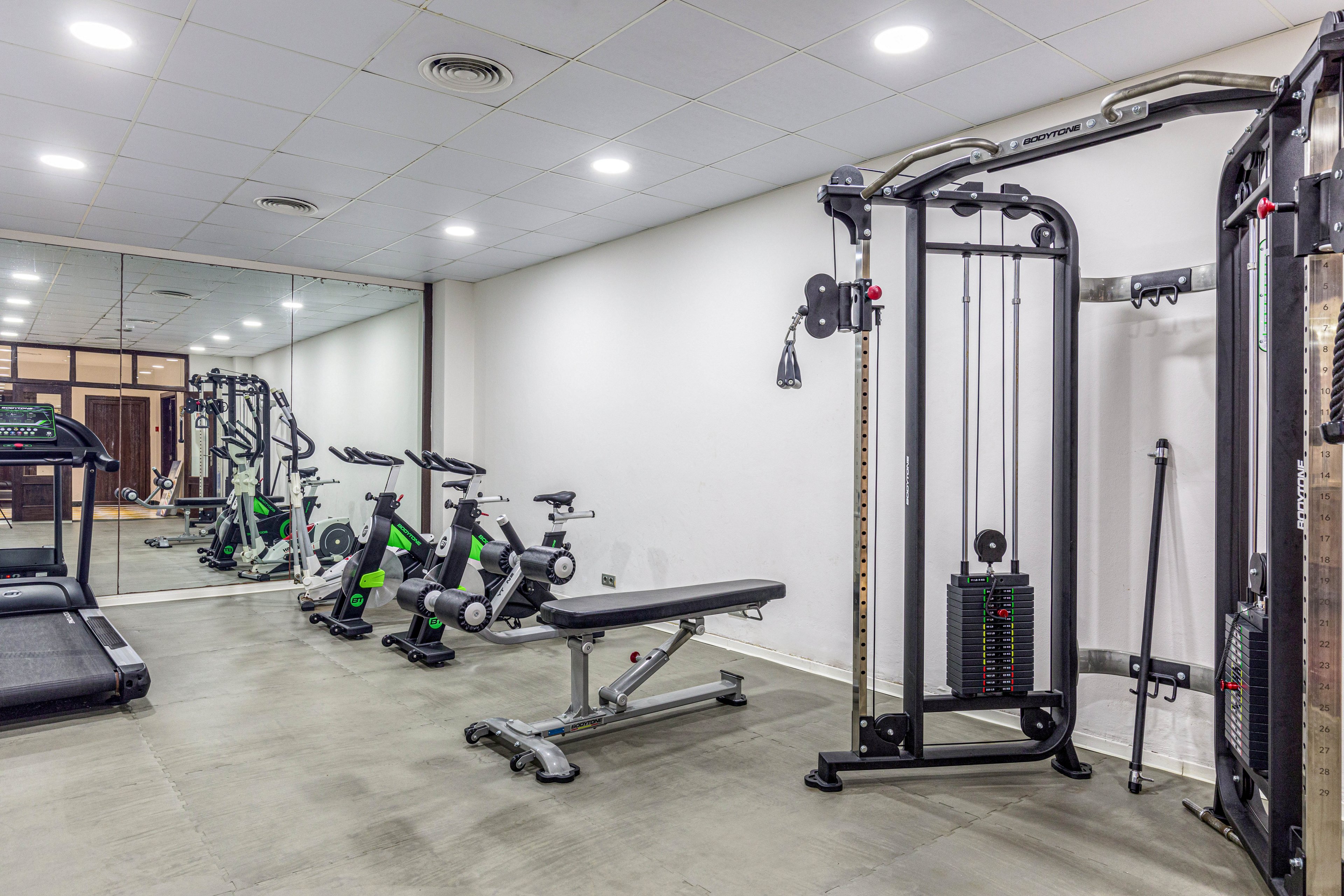 A clean, modern gym room features various exercise machines neatly arranged along the walls. A cable machine and adjustable weight bench stand in the foreground on a polished concrete floor. In the background, treadmills and stationary bikes face a large wall mirror that visually doubles the space. Bright, even ceiling lighting and white walls create a minimalist, professional workout environment.