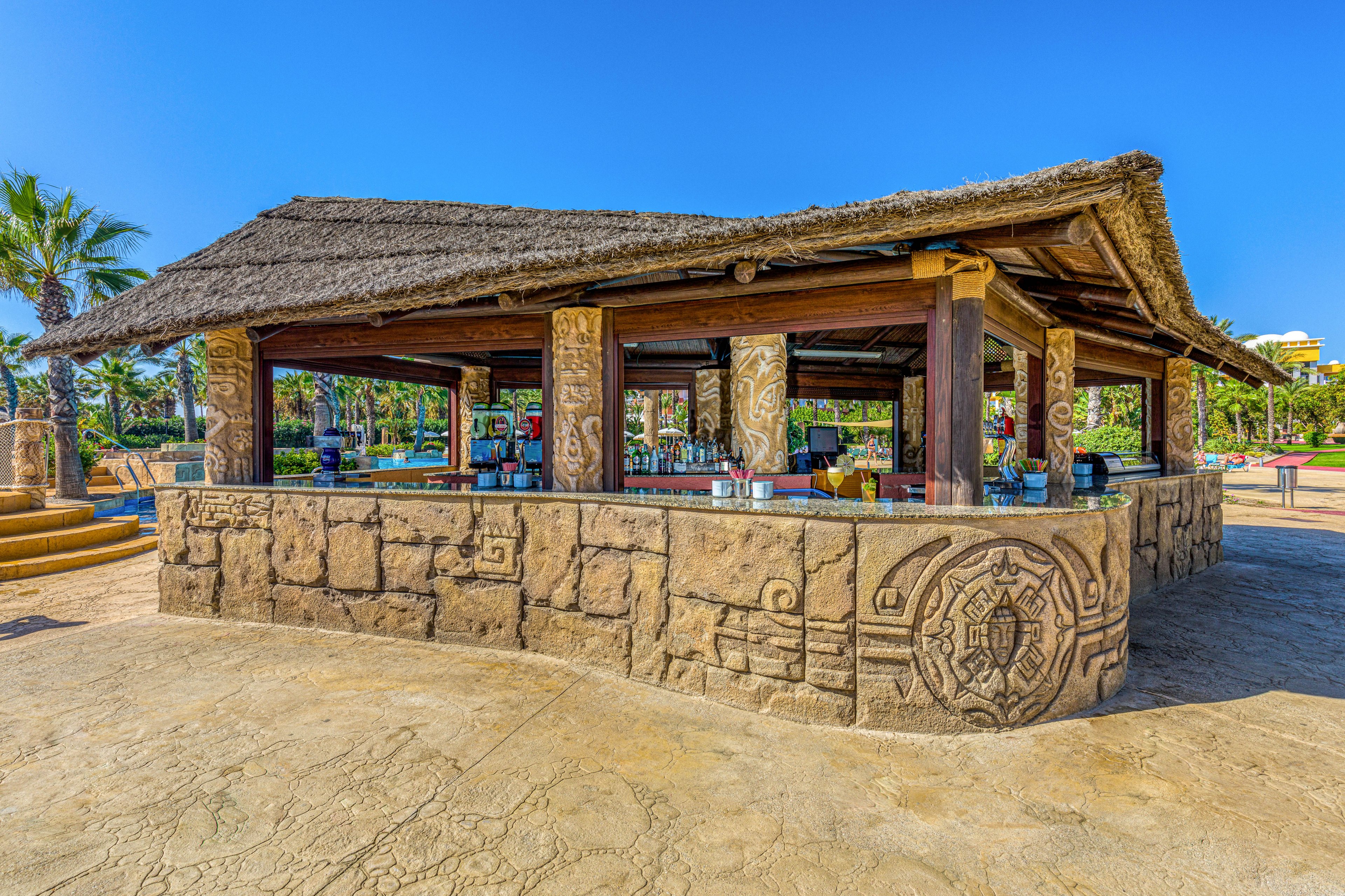 An open-air bar with a thatched roof stands in a sunny resort setting. The bar is built from decorative stone with carved patterns and columns, giving it a tropical, rustic feel. Beverage dispensers and bar equipment are visible inside the shaded interior. Palm trees, manicured greenery, and clear blue sky surround the structure, suggesting a warm vacation destination.