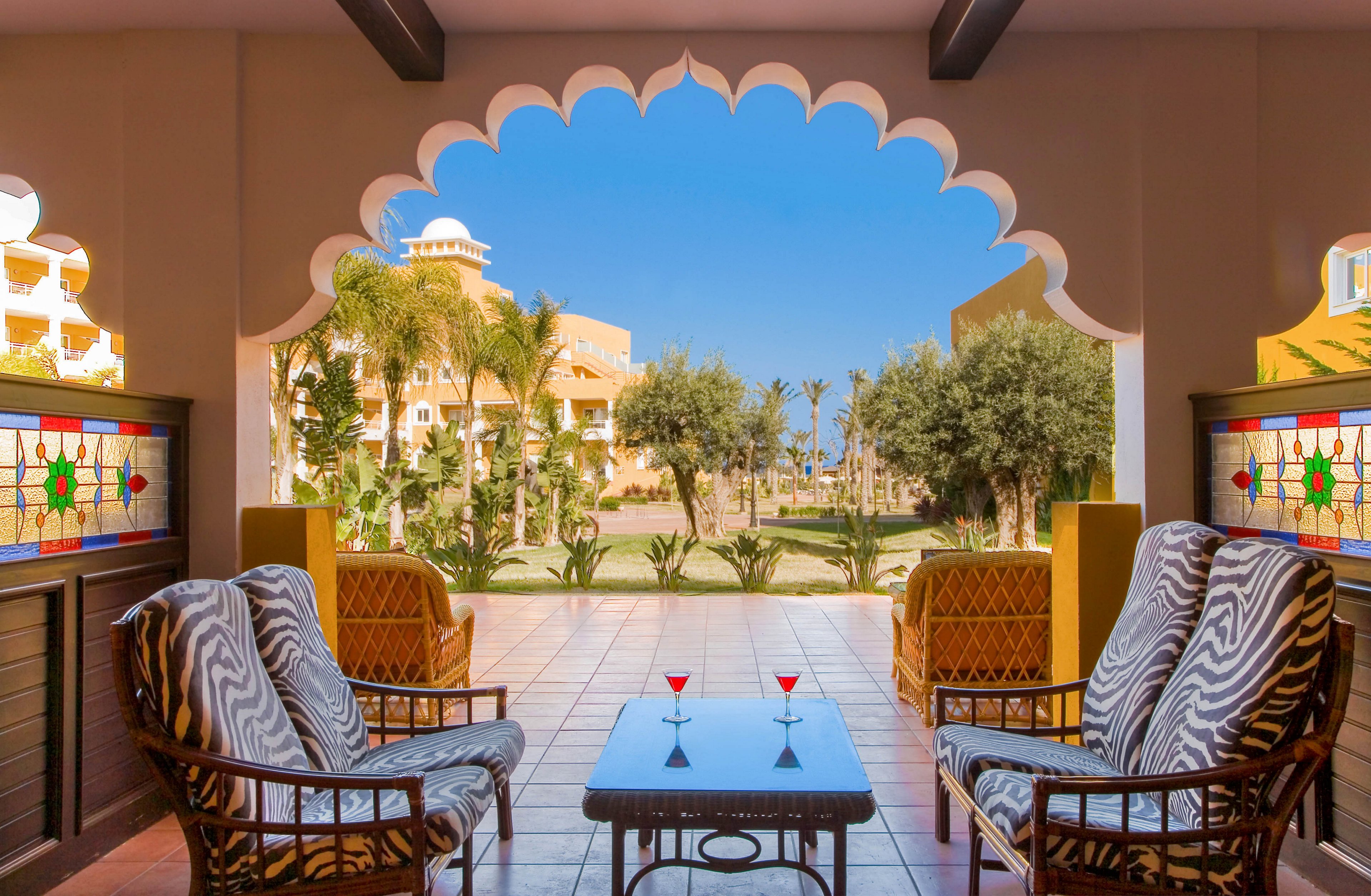 An inviting resort terrace features cushioned wicker chairs arranged around a small glass-top table with colorful drinks. The scene opens through an ornate arched frame onto a lush garden with palm trees and warm-toned buildings in the background. Stained glass panels and patterned upholstery add decorative detail to the cozy seating area. Clear blue sky and bright sunlight create a relaxed, vacation atmosphere.