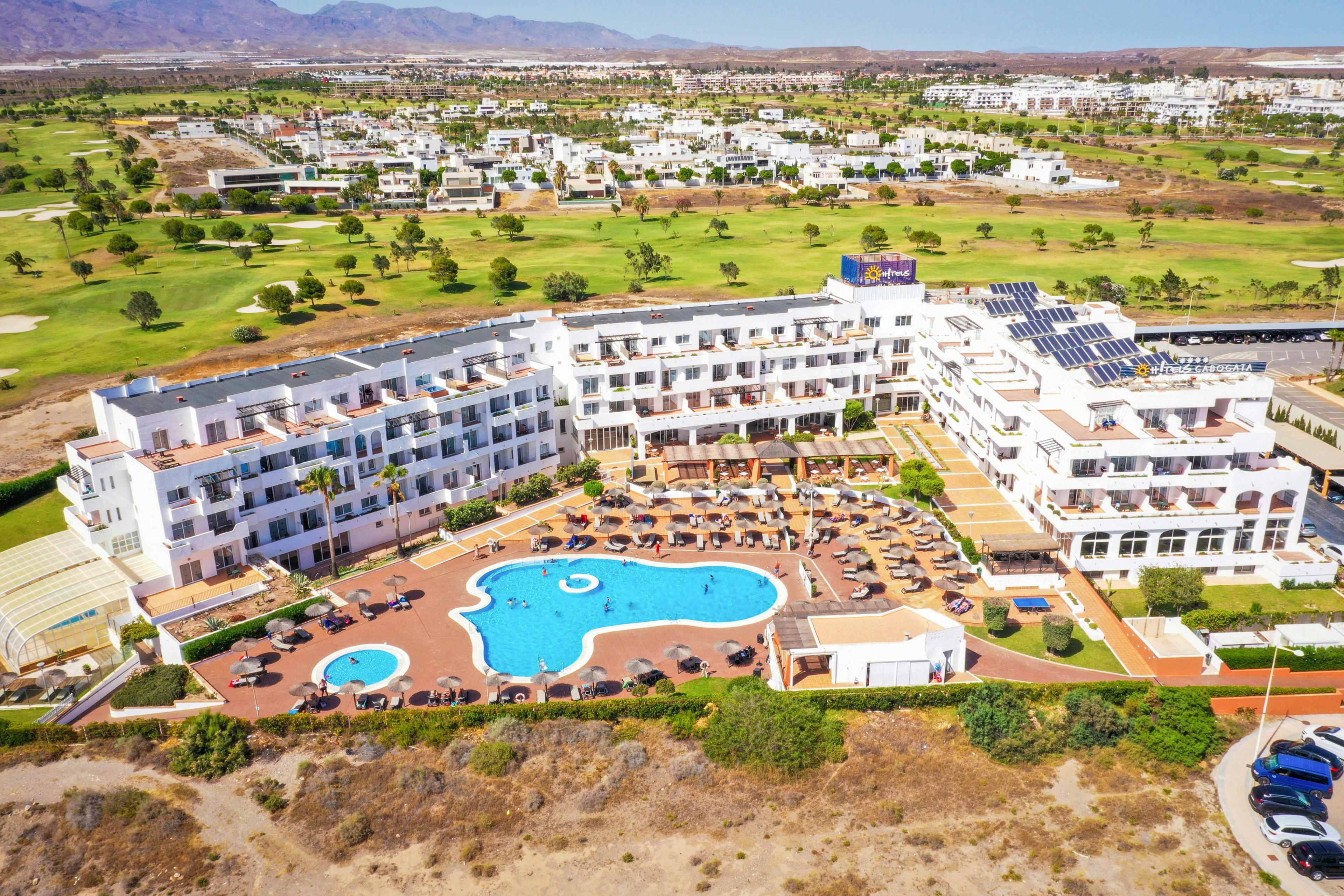 A large resort hotel complex is captured from an aerial perspective, showcasing multiple white buildings arranged around a central courtyard. In the middle, a bright blue outdoor swimming pool with curved shapes is surrounded by sun loungers and umbrellas. The property is set on the edge of a dry, sandy foreground with parked cars visible to one side. Beyond the hotel, expansive green golf course fairways and scattered trees stretch toward distant mountains under a clear sky.