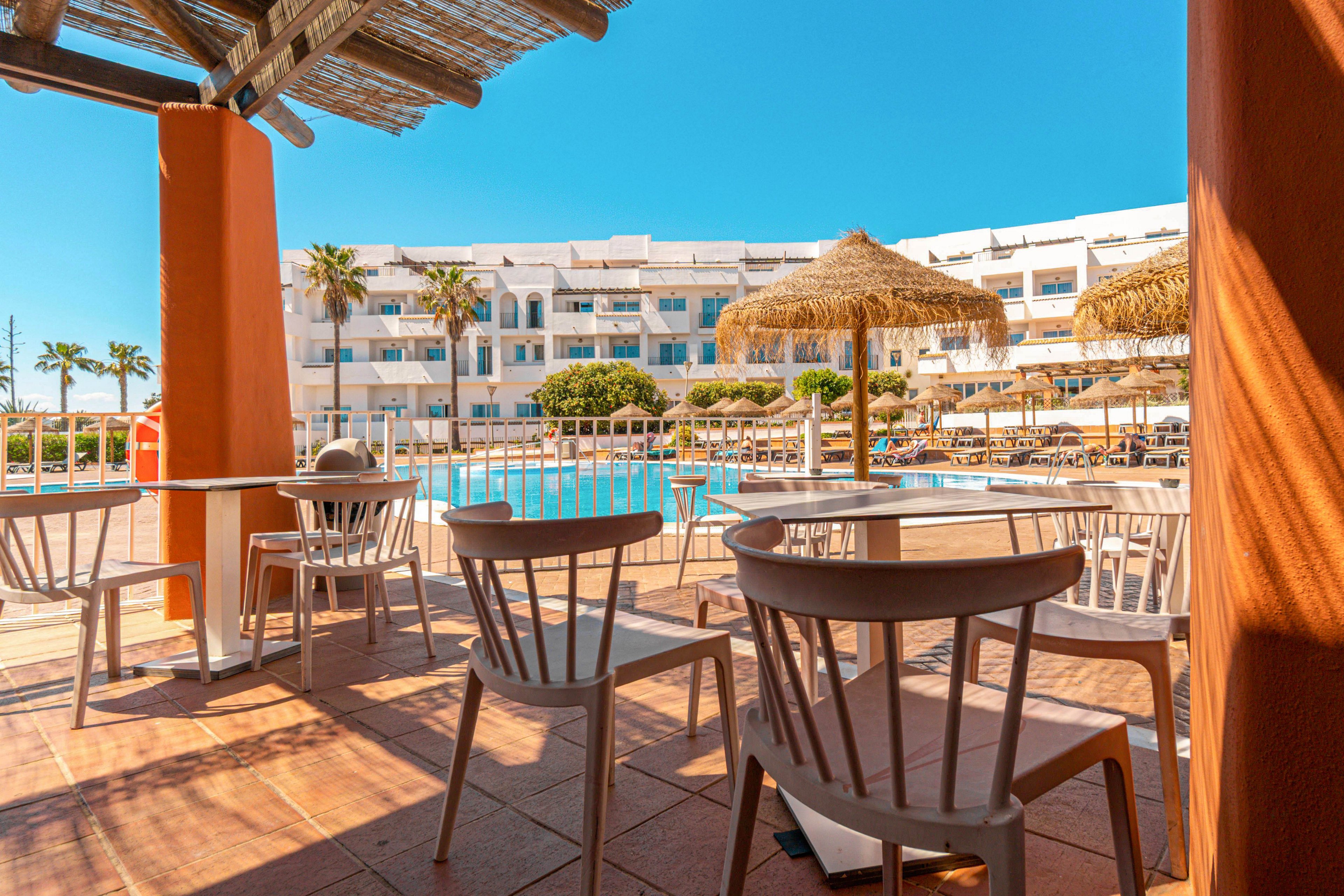 An outdoor resort terrace overlooks a bright blue swimming pool on a sunny day. White chairs and tables sit in the shade of a wooden pergola, facing the pool area. Straw parasols and sun loungers line the pool deck, with a white multi-story hotel building in the background. Palm trees and clear blue sky enhance the relaxed vacation atmosphere.