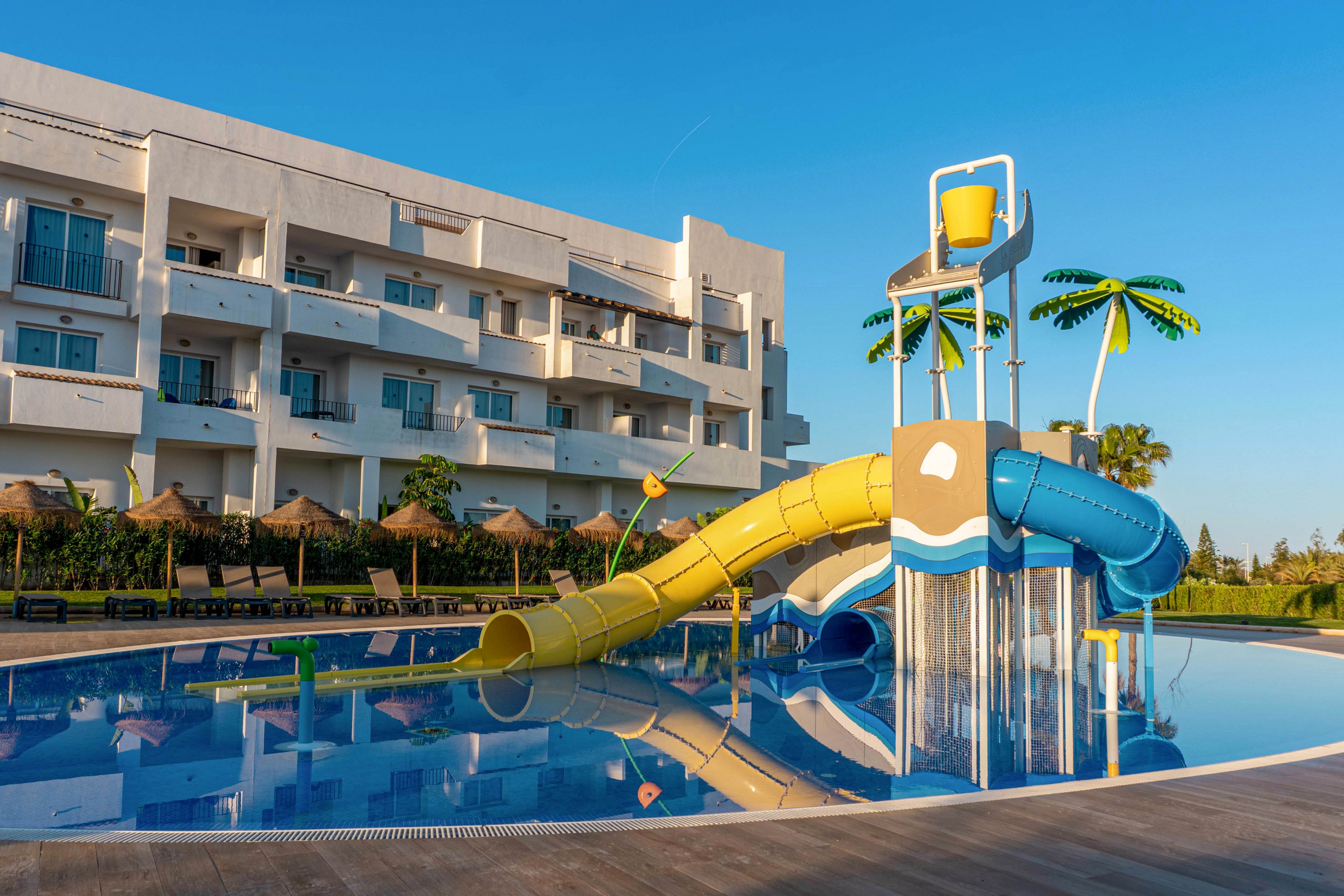 A colorful waterslide playground stands in a shallow resort pool in front of a modern white hotel building. The structure features yellow and blue slides, a large tipping bucket, and decorative palm tree elements. Sun loungers and straw parasols line the poolside area, suggesting a relaxing vacation setting. The clear blue sky and calm water reflections create a bright, family-friendly atmosphere.