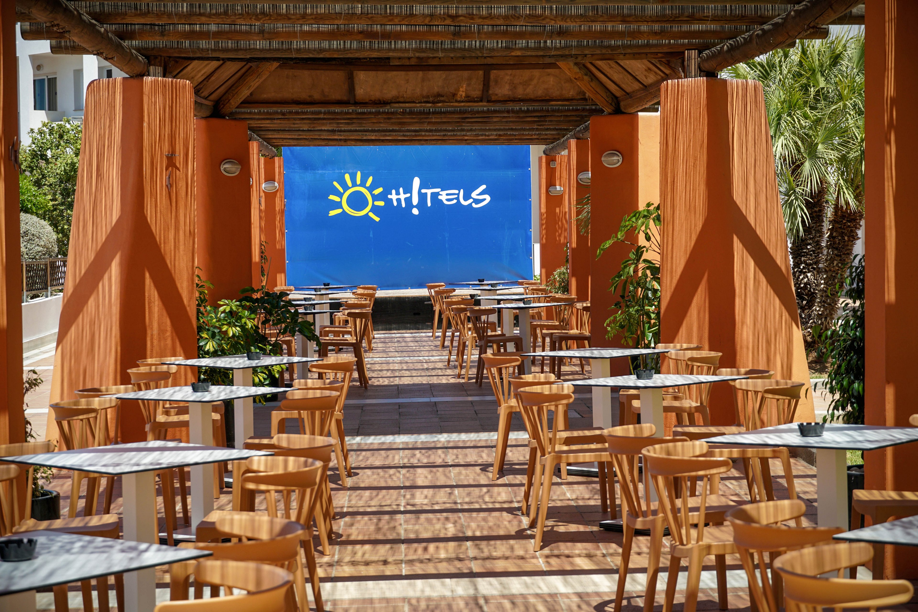 An open-air hotel terrace café features rows of wooden chairs and small tables arranged under a shaded pergola. Warm orange columns frame the walkway, leading the eye toward a bright blue wall at the far end. A stylized sun graphic and the word "HOTELS" are clearly visible on the blue backdrop. Lush green plants and palm trees surround the space, creating a relaxed vacation atmosphere.