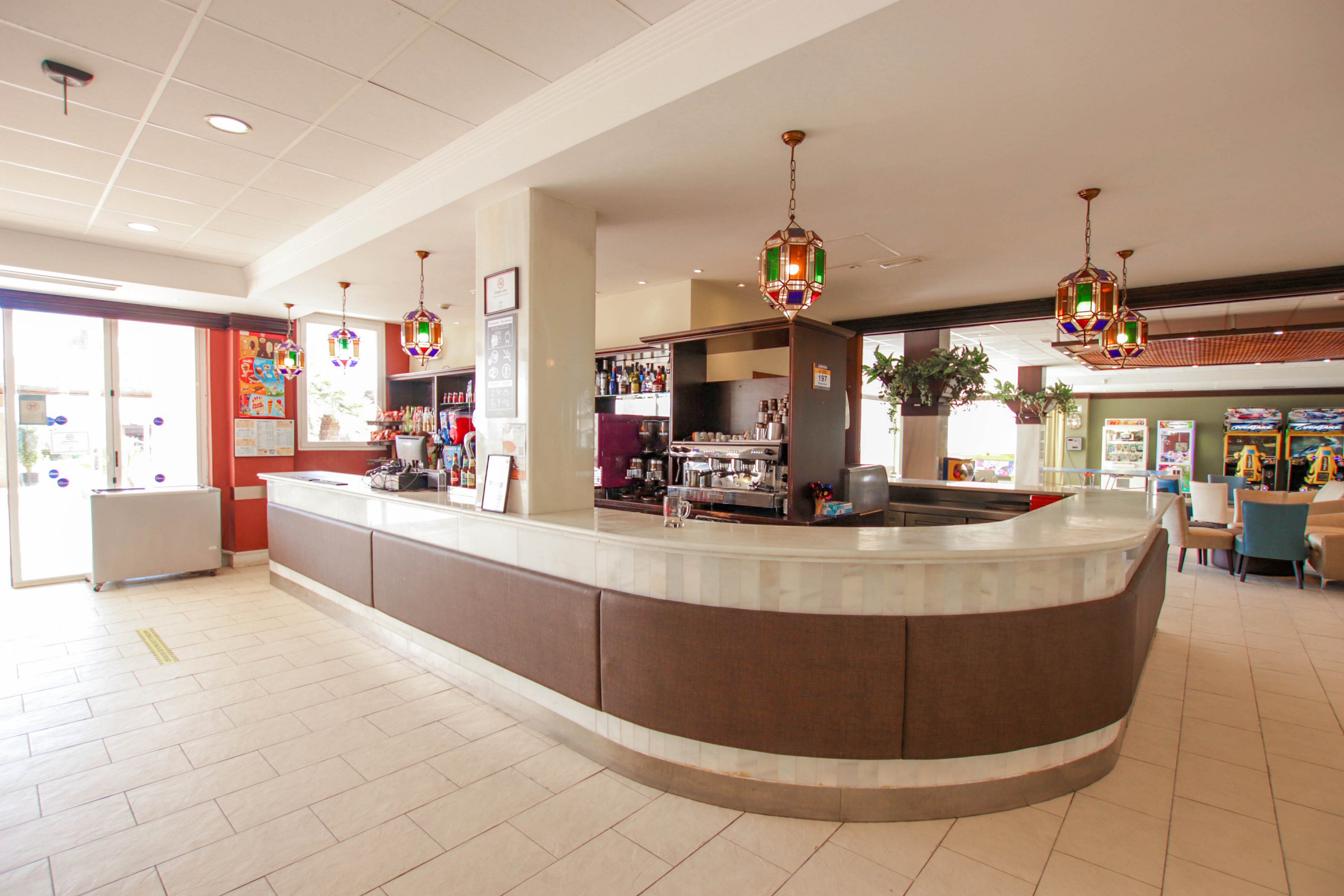 A spacious bar area features a long curved counter with a light stone top and brown paneling. Colorful pendant lights hang from the ceiling, illuminating the bar shelves stocked with bottles and glasses. Large windows on the left bring in natural daylight, while seating and lounge areas are visible in the background. The clean tiled floor and neutral walls create an airy, welcoming atmosphere suitable for guests to relax and socialize.