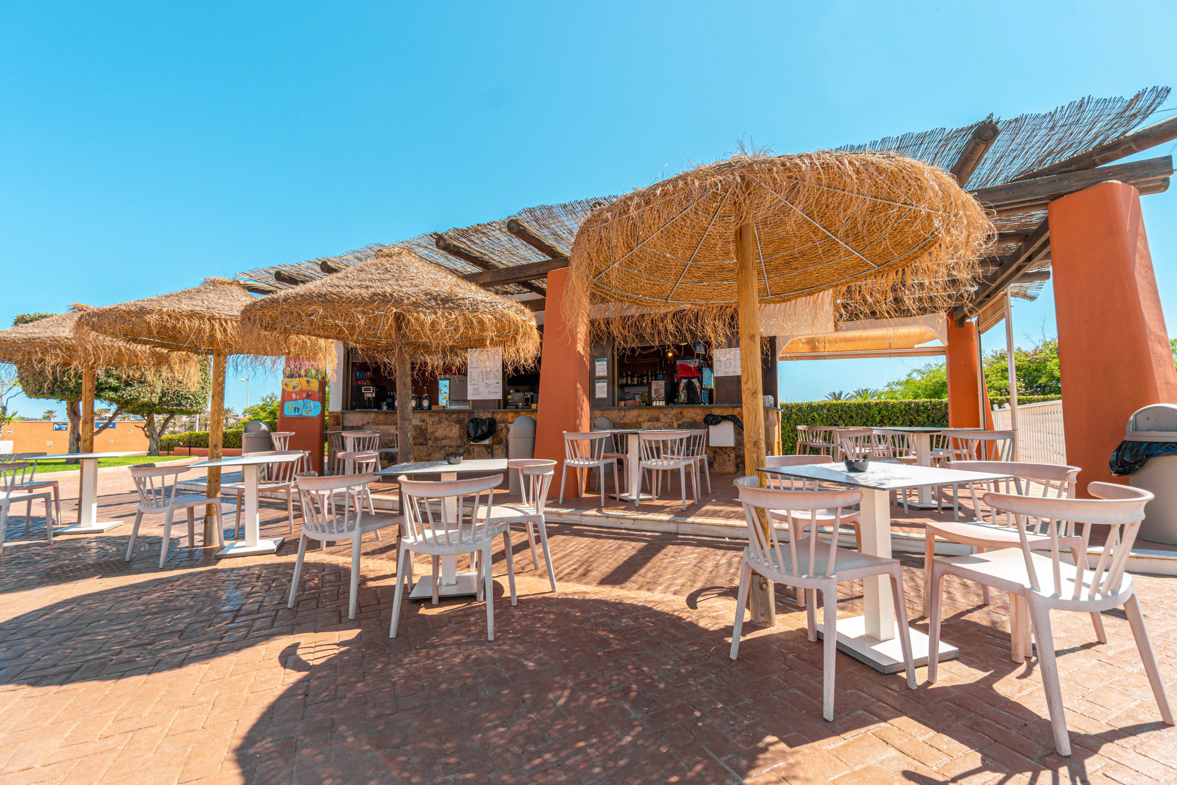An open-air bar features several straw parasol roofs shading a counter and seating area. White tables and chairs are arranged on a sunlit stone patio in front of the bar. The scene is bright and clear under a vivid blue sky, suggesting a warm summer day. Greenery and distant landscape elements frame the relaxed coastal atmosphere.
