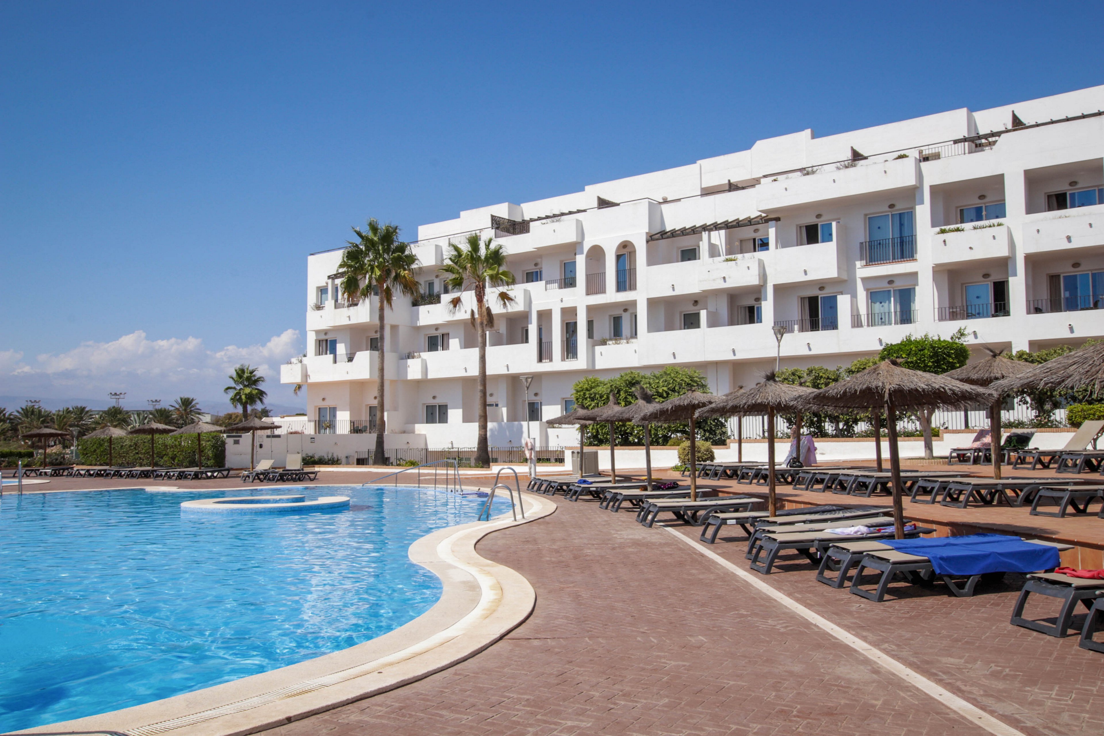 A large outdoor swimming pool curves alongside a modern white resort hotel. Numerous sun loungers and straw parasols line the poolside terrace, suggesting a relaxing vacation setting. Palm trees and manicured shrubs add a tropical touch under a clear blue sky. The scene appears calm and uncrowded, emphasizing leisure and warm weather holidays.