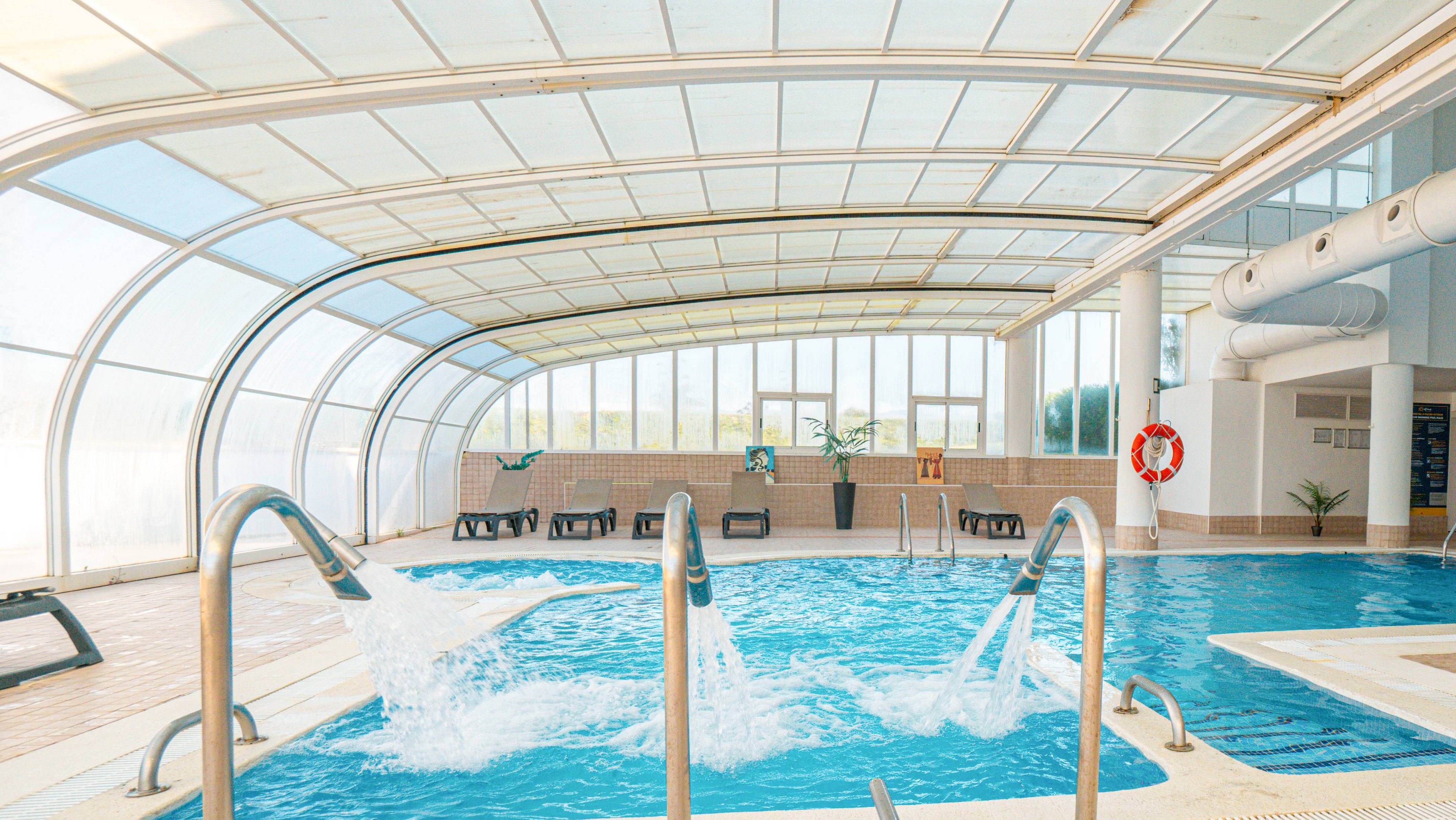 An indoor swimming pool features strong water jets cascading from stainless steel pipes into the clear blue water. The pool is housed under a bright, curved glass and metal roof that lets in natural light. Lounge chairs line the far side of the pool, suggesting a relaxing spa or wellness setting. Safety elements like a life preserver and railings are visible around the pool area.