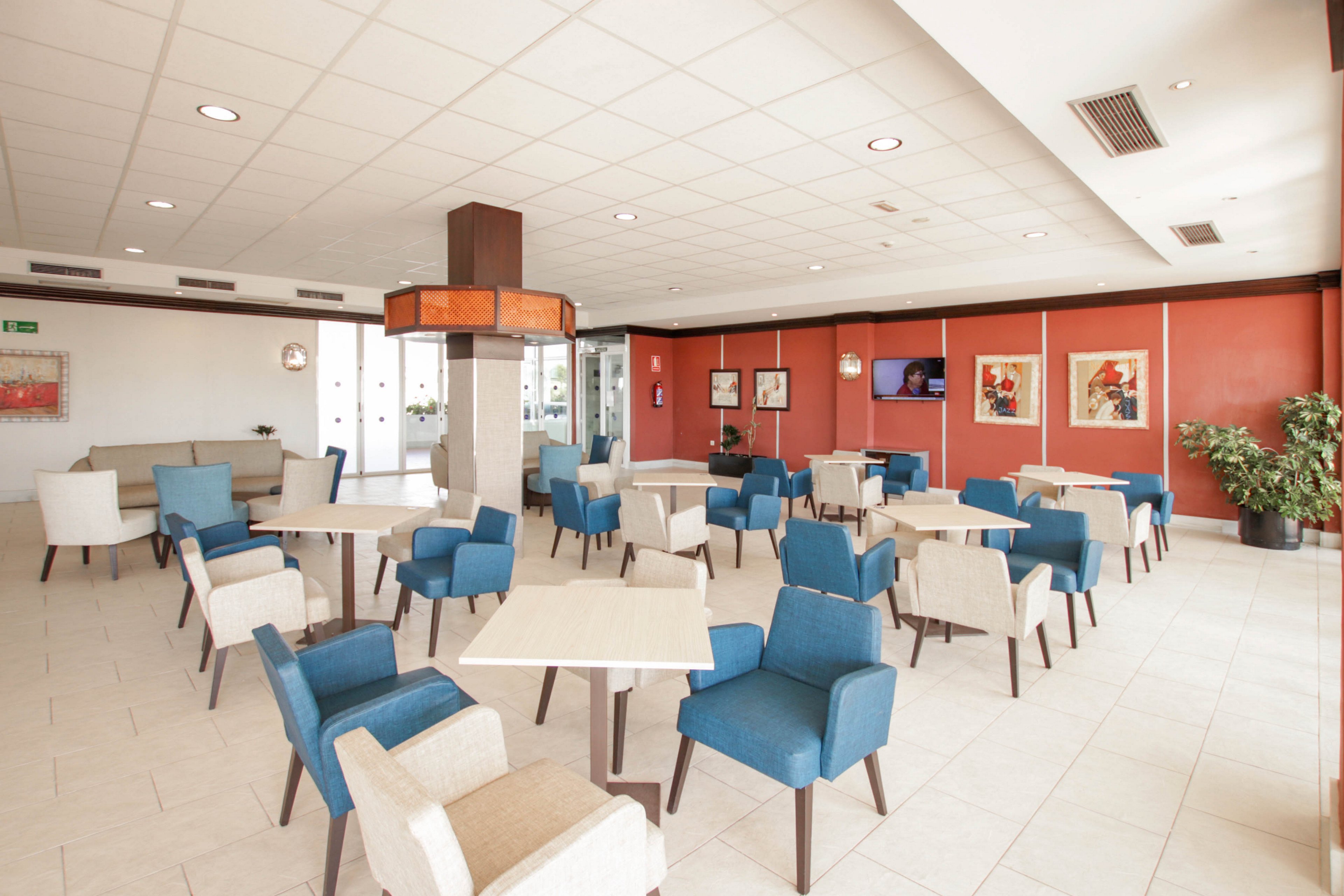 Spacious, modern lounge area with multiple square tables and upholstered chairs in blue and beige tones. The room is empty, with no people present, and features a tiled floor and a white grid ceiling with recessed lighting. Large windows at the back let in natural light, while framed artwork decorates the red accent walls. A television screen and potted plant add to the contemporary, welcoming atmosphere.