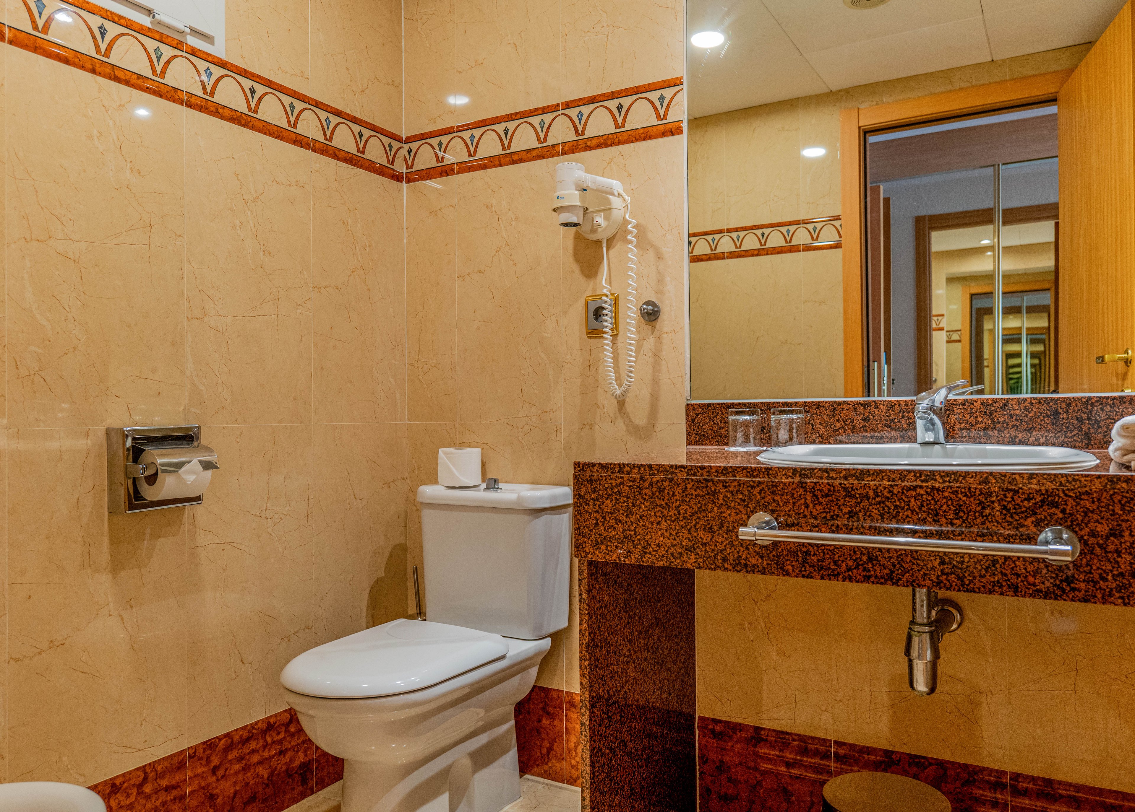 A clean, modern bathroom features a white toilet and a granite-style vanity countertop. The space is finished with beige and brown tiled walls accented by a decorative border. A large mirror above the sink reflects the open wooden door and part of the room beyond. A mounted hair dryer, metal towel bar, and neatly arranged toiletries emphasize a functional hotel-style setting.