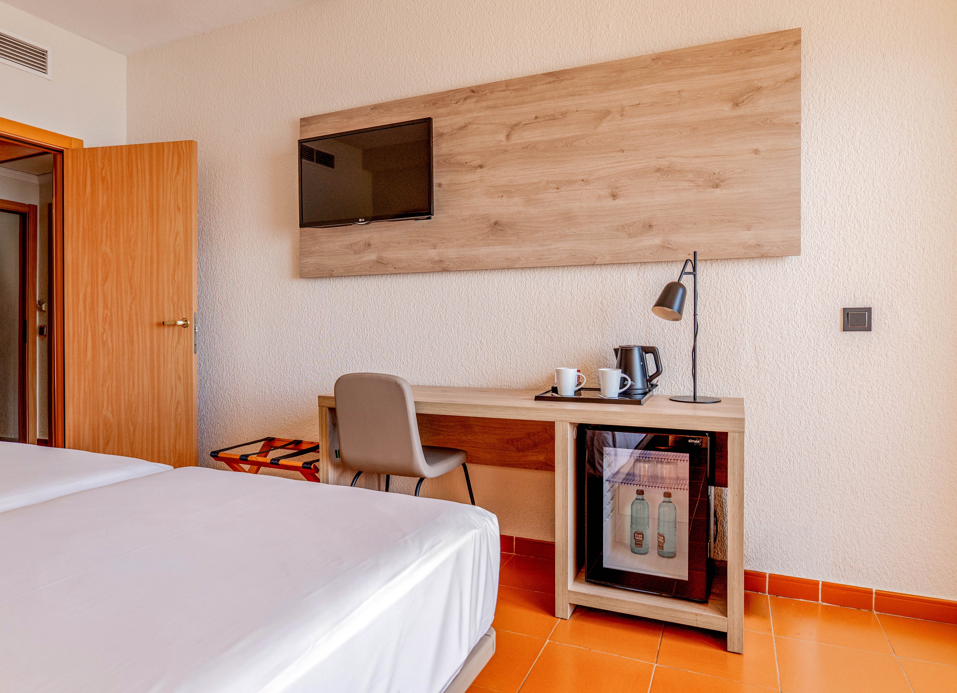 A bright hotel room features a neatly made bed in the foreground and a work desk against the wall. Above the desk, a flat-screen television is mounted on a wooden panel. The desk holds a desk lamp, cups, and a kettle, while a small minibar fridge with drinks inside sits beneath. An open wooden door and orange floor tiles add warmth to the simple, modern interior.