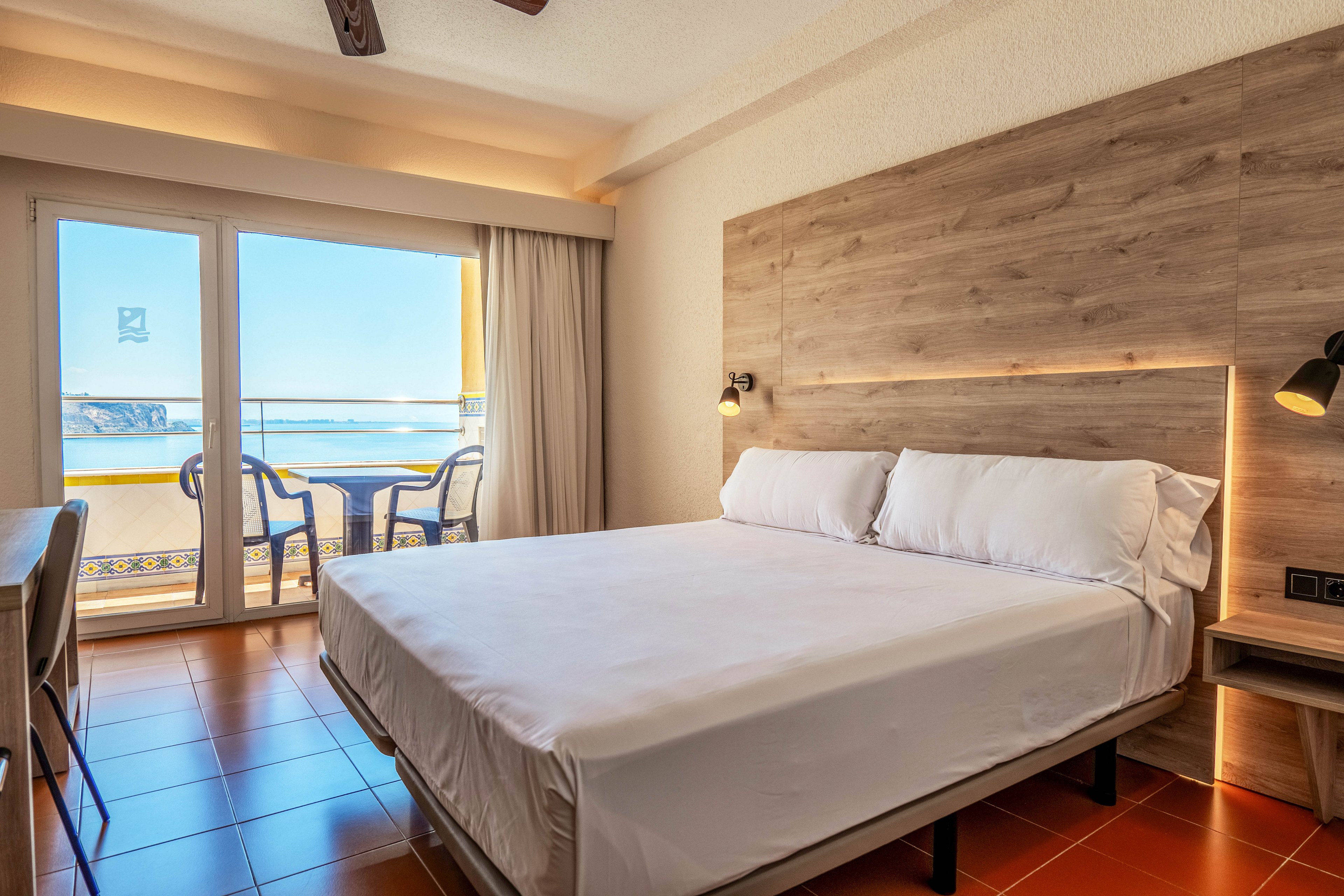 A neatly made double bed with white linens sits in a modern hotel room featuring a wooden headboard and warm lighting. Large glass doors open onto a balcony with two chairs and a small table overlooking the sea and distant cliffs. Terracotta floor tiles and neutral walls create a clean, inviting atmosphere. The space feels bright and airy, ideal for a relaxing coastal getaway.