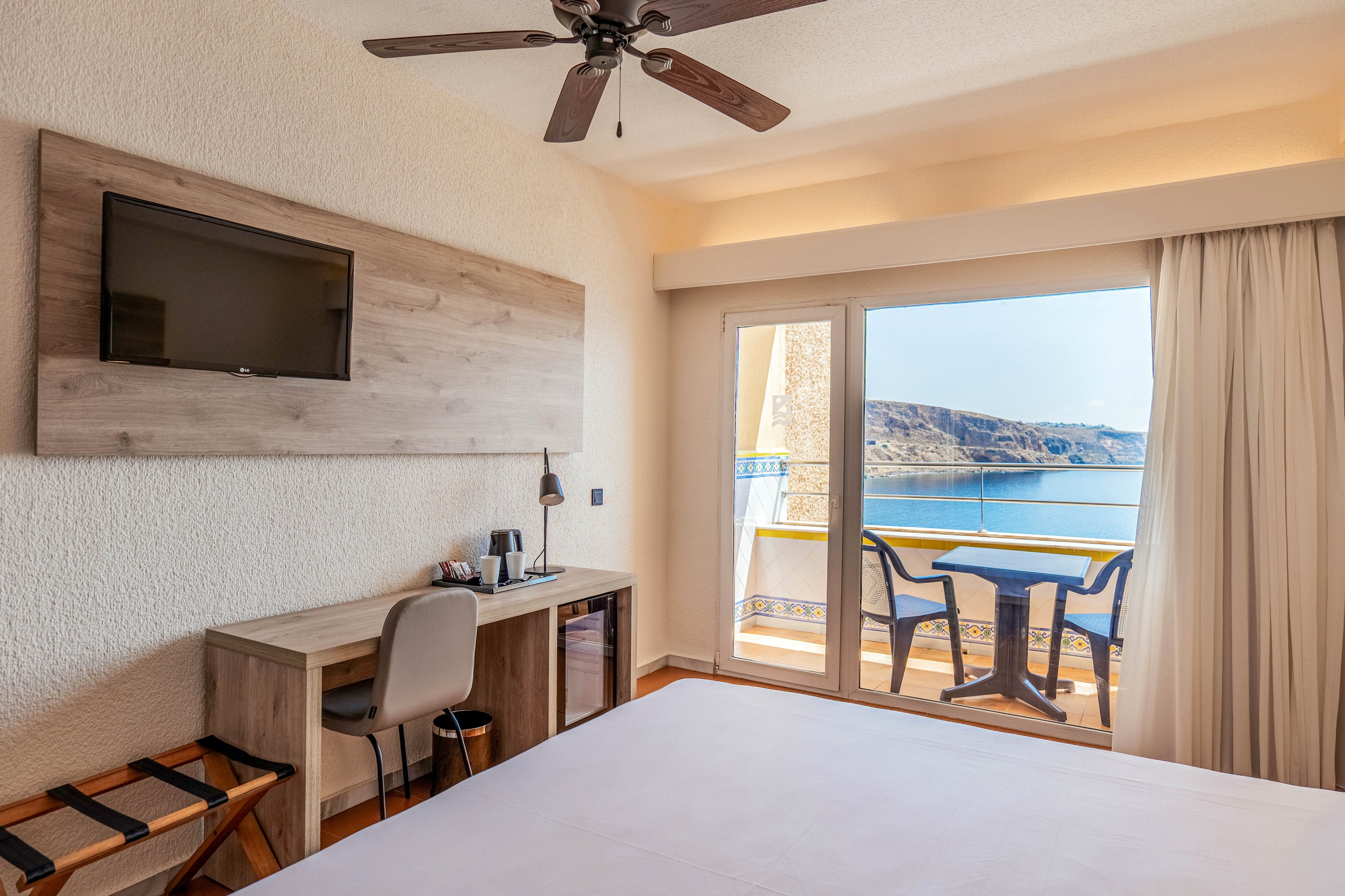 A bright hotel room features a neatly made bed, a wall-mounted television, and a compact work desk with a chair. Sliding glass doors open onto a private balcony with two chairs and a small table overlooking a calm sea and distant coastline. Warm neutral tones, wooden accents, and a ceiling fan create a relaxed, contemporary atmosphere. Natural daylight fills the space, highlighting the inviting view and comfortable furnishings.