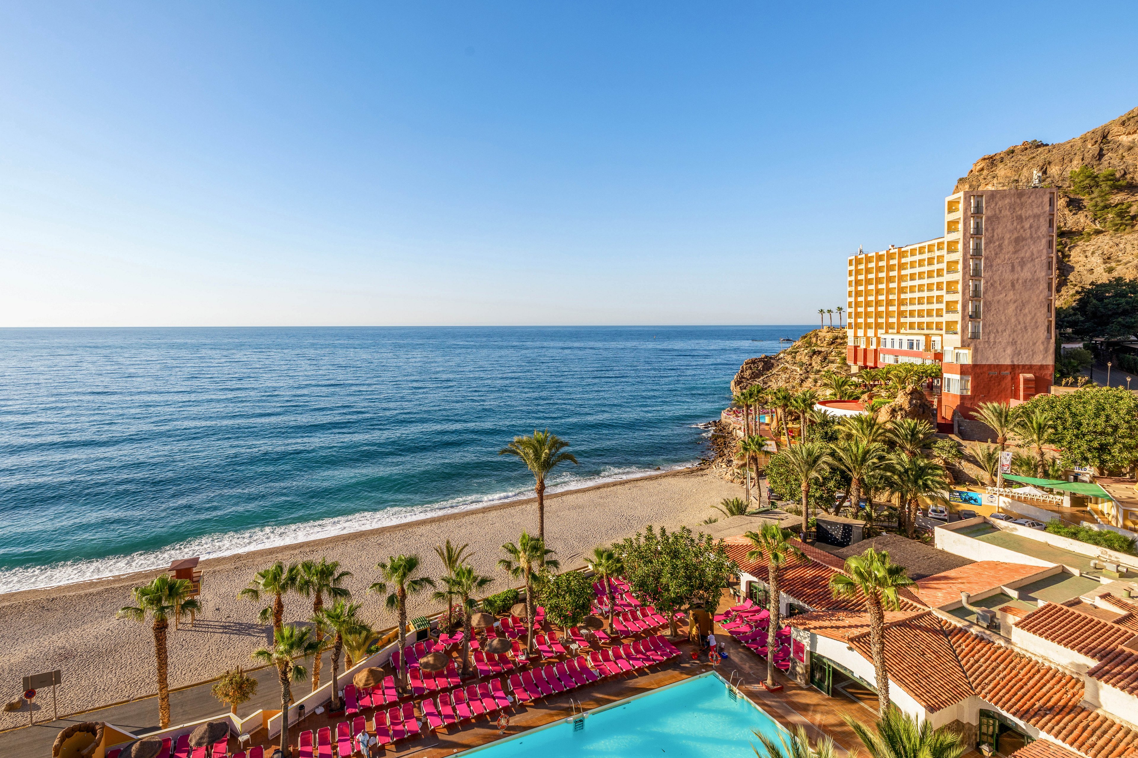 A coastal resort overlooks a wide sandy beach and calm blue sea under a clear sky. In the foreground, a bright turquoise swimming pool is surrounded by rows of pink lounge chairs and palm trees. A multi-story hotel building stands on the rocky hillside to the right, facing the ocean. The scene suggests a sunny, relaxing vacation destination with direct beach access and ocean views.