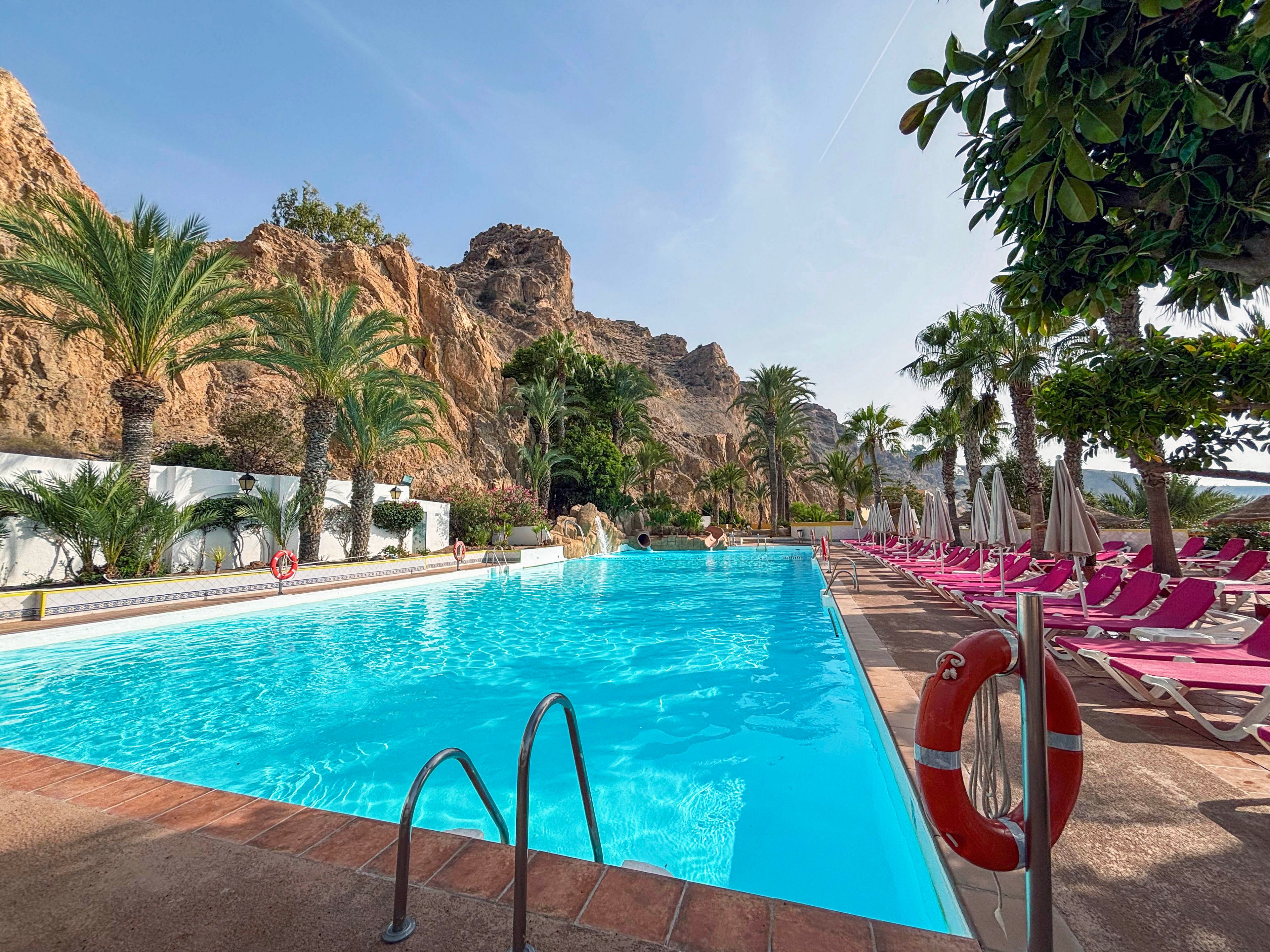 A large outdoor swimming pool stretches alongside a row of pink lounge chairs and umbrellas at a resort. Rocky cliffs and palm trees rise dramatically in the background, creating a striking contrast with the clear blue water. Metal pool ladders and red-and-white life preservers frame the foreground, emphasizing safety and access. The bright daylight and clear sky suggest a warm, relaxing vacation atmosphere.