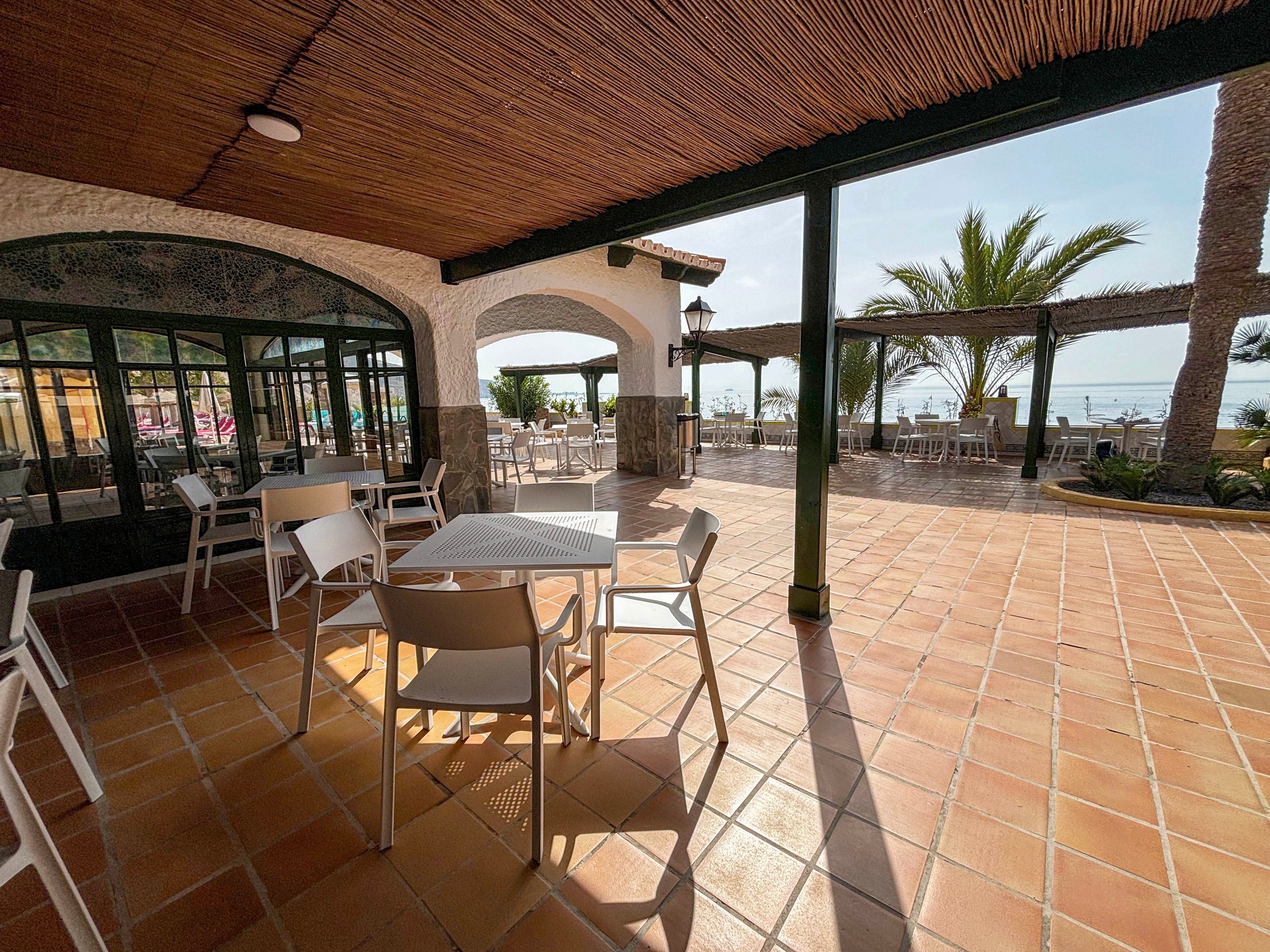An open-air seaside terrace features several empty white tables and chairs under a shaded pergola. The tiled floor extends toward an outdoor seating area with views of the ocean in the distance. Palm trees and wooden beams frame the bright, sunny coastal setting. Strong sunlight casts long shadows, emphasizing the relaxed, vacation atmosphere.