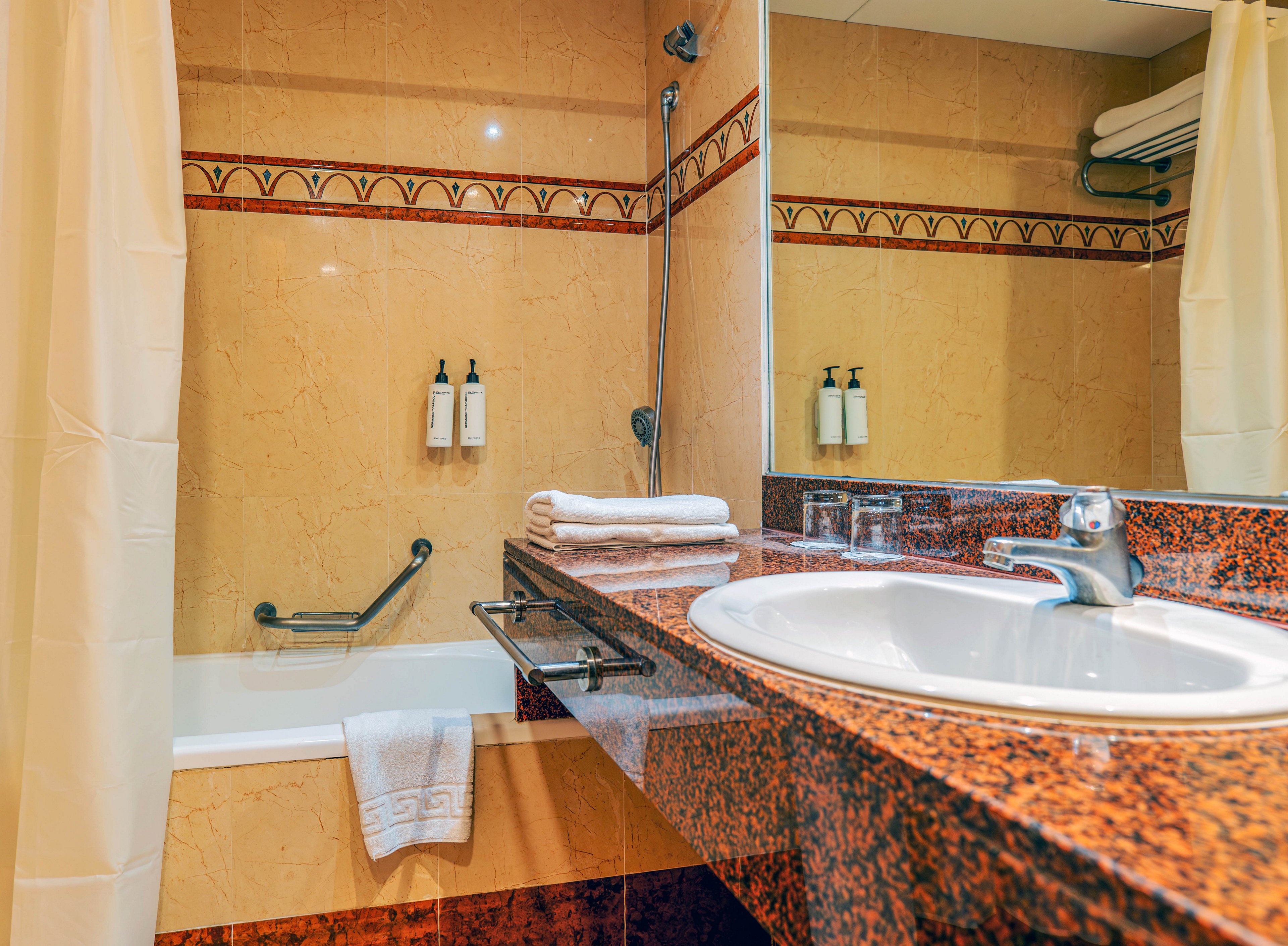 A clean hotel bathroom features a white sink set into a polished stone vanity counter. A bathtub with a mounted showerhead and safety grab bar sits beside it, framed by cream-colored shower curtains. Fresh white towels are neatly folded on the counter and hanging over the tub edge, with two wall-mounted toiletry dispensers above. Warm beige and brown tiles and a large mirror create a cozy, inviting atmosphere.