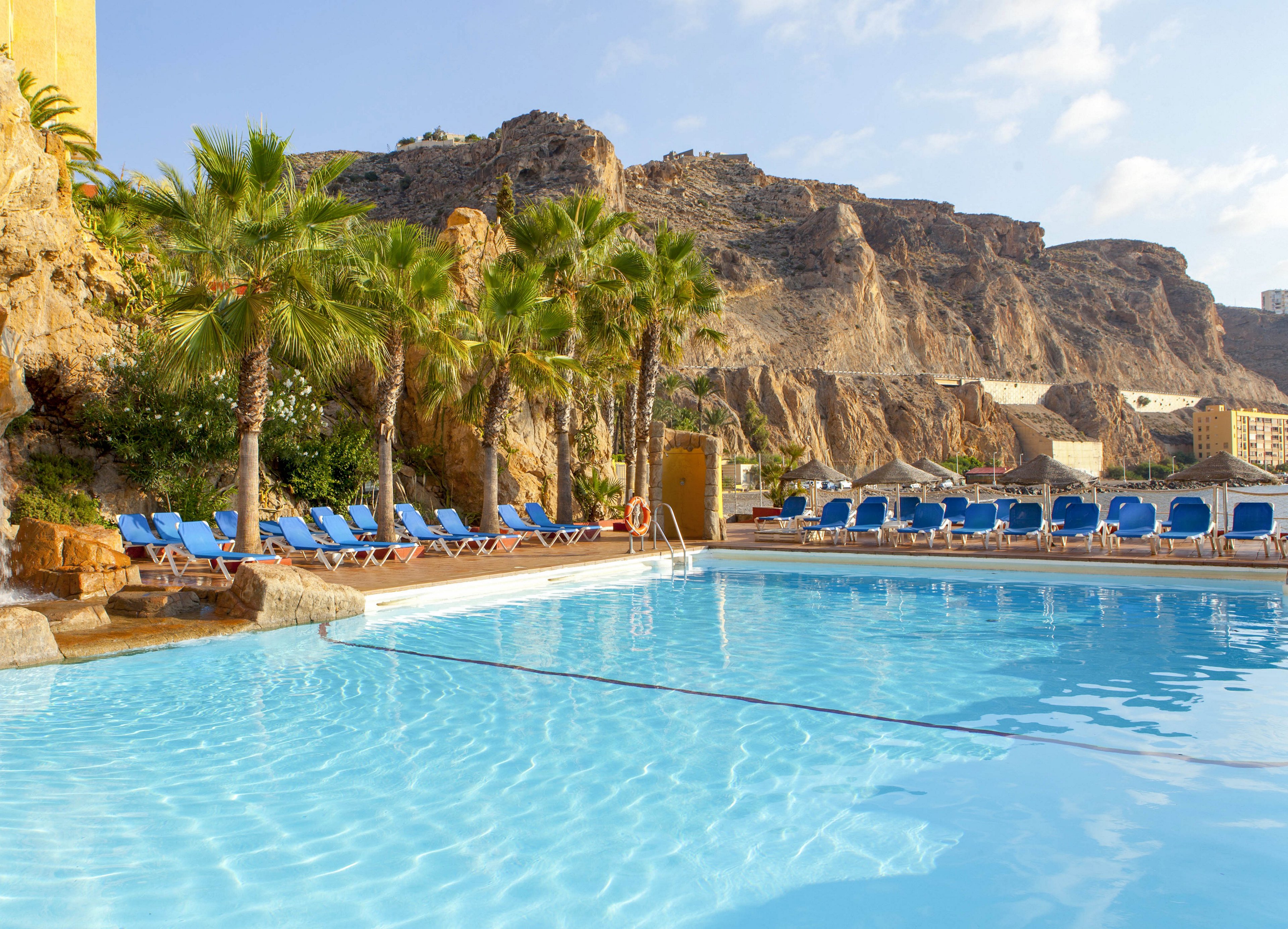 An outdoor resort swimming pool glistens under bright sunlight, surrounded by rows of blue lounge chairs. Tall palm trees line the poolside, adding a tropical feel to the scene. Rocky mountains rise in the background, with buildings nestled into the hillside. The clear blue water and open sky create a relaxing vacation atmosphere.