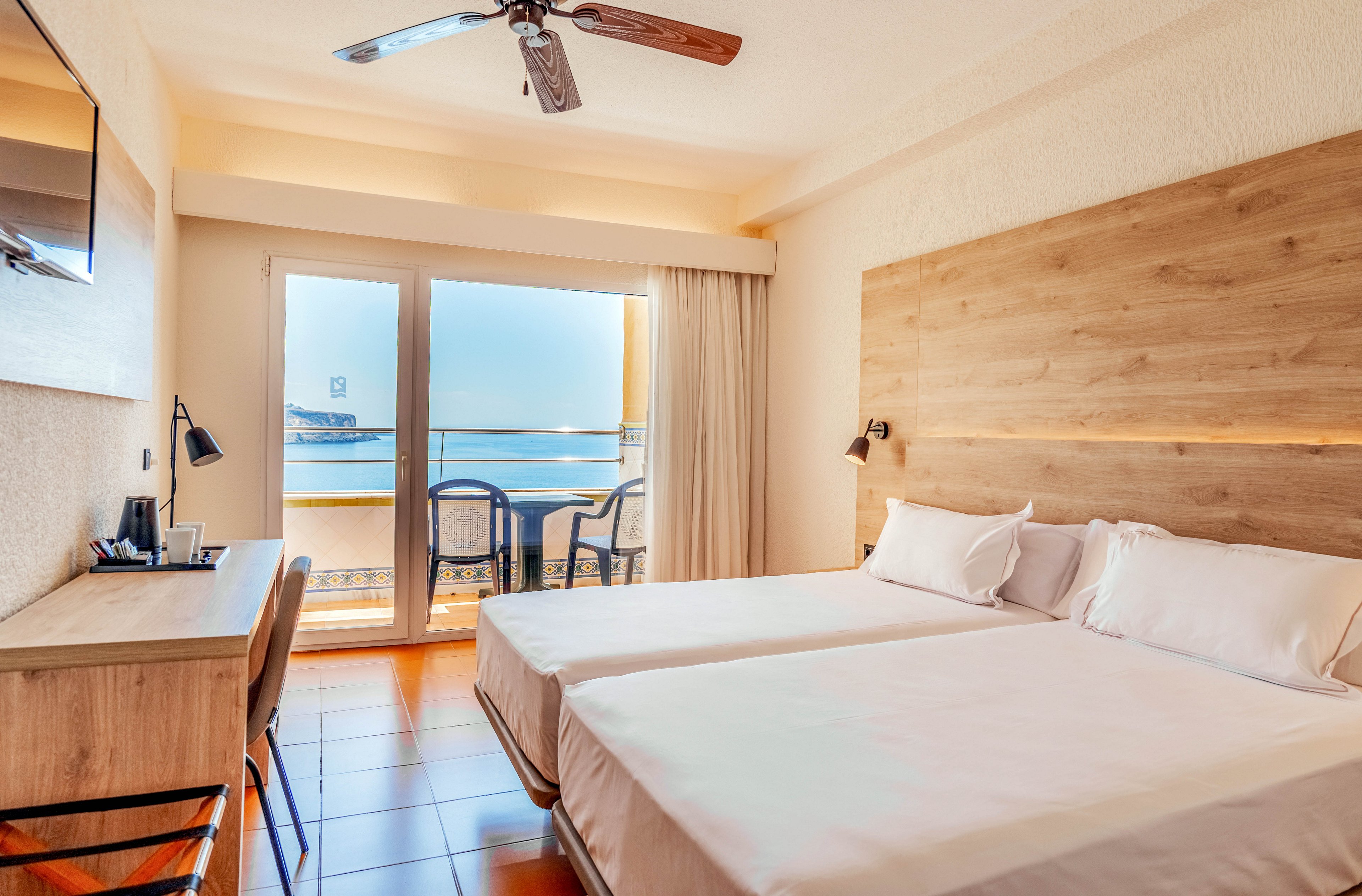 A bright hotel room features two neatly made twin beds with white linens and a wooden headboard. A desk with a chair, wall-mounted television, and bedside lamps complete the modern furnishings. Large glass doors open onto a balcony with two chairs, offering a scenic view of the sea and distant coastline. Warm lighting and neutral tones create a calm, inviting atmosphere suitable for vacation or business travel.