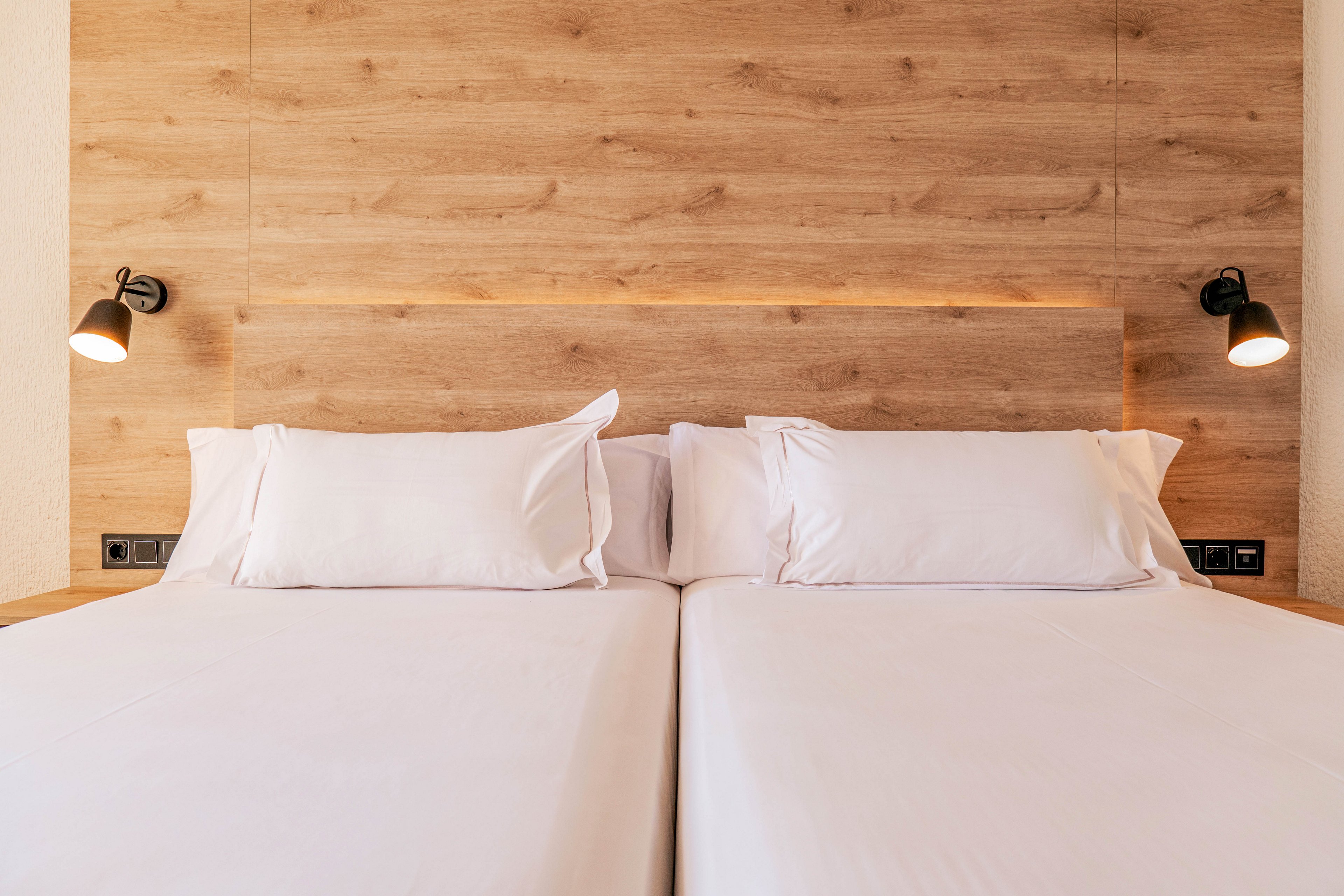 Two neatly made twin beds are placed side by side, topped with crisp white sheets and pillows. The beds sit against a light wooden headboard that adds warmth and texture to the room. Two black wall-mounted reading lamps flank the headboard, casting soft light on the sleeping area. The overall scene suggests a clean, modern hotel or guest room designed for comfort and simplicity.