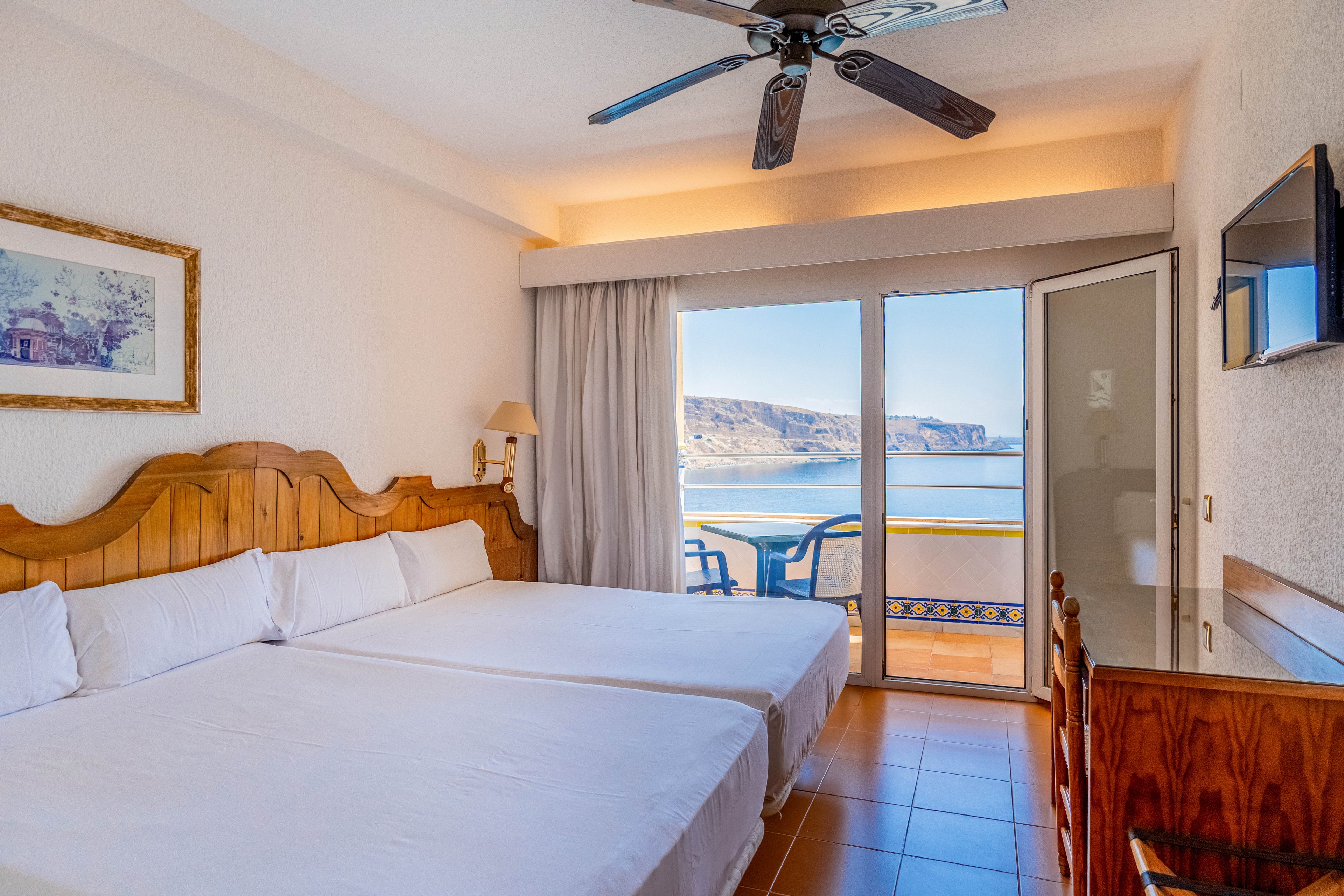 A bright hotel room features two neatly made twin beds with wooden headboards. The space includes a wall-mounted television, a desk, and a ceiling fan above. Large glass doors open onto a balcony with blue chairs, revealing a scenic ocean and cliffside view. Warm natural light fills the room, creating an inviting and relaxing atmosphere.