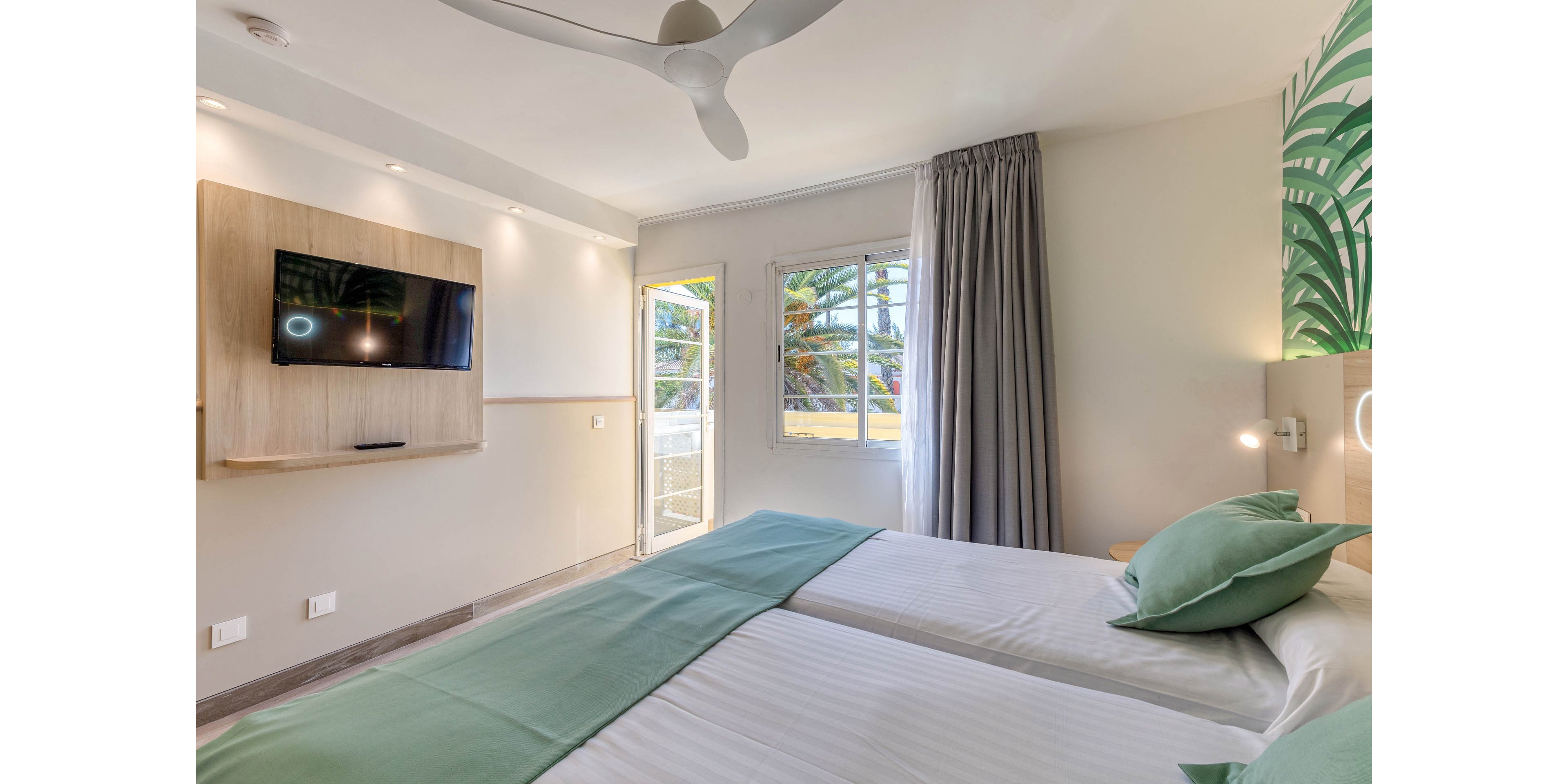 Contemporary hotel room featuring two neatly made twin beds with mint green accents. A wall-mounted flat-screen TV faces the beds, and a ceiling fan hangs above. Natural light enters through a window and glass door leading to a balcony with palm trees outside. Soft neutral walls, a tropical leaf mural, and simple bedside lighting create a calm, inviting atmosphere.