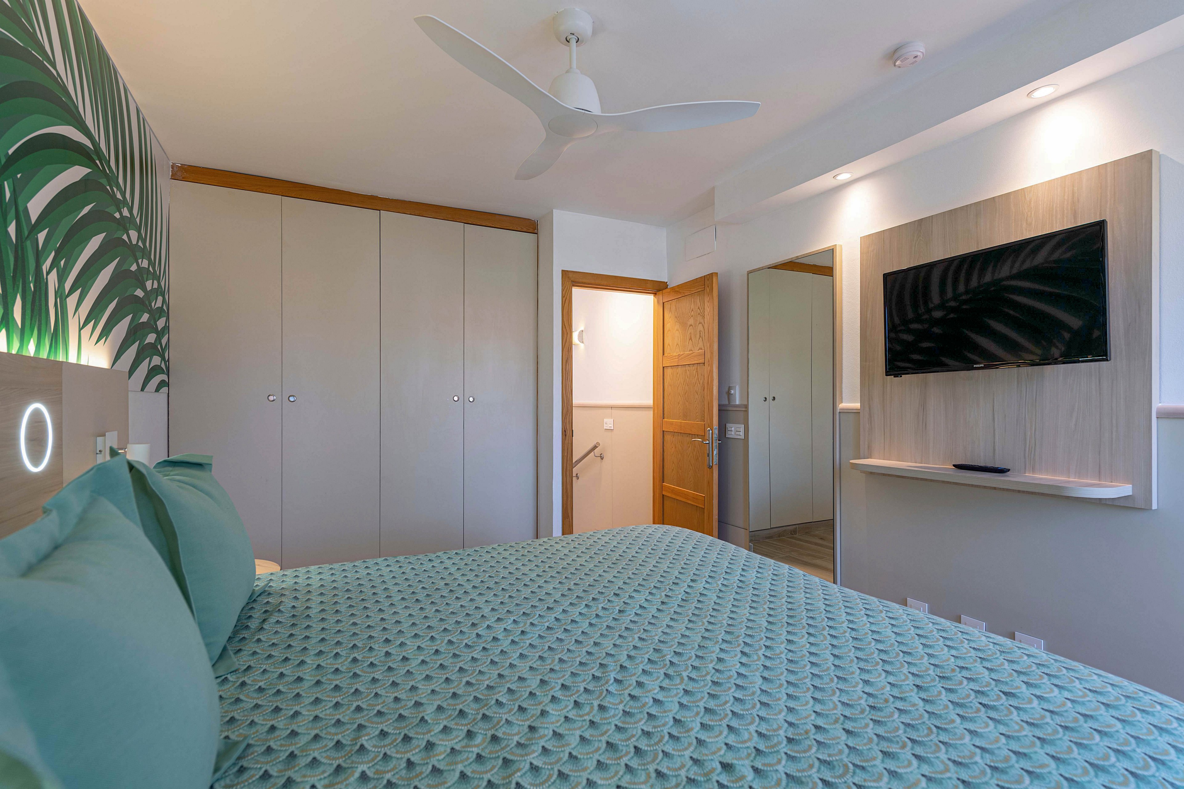 A contemporary bedroom features a neatly made bed with textured teal bedding and matching pillows. Built-in wardrobes with minimalist doors line one wall, while a wooden door opens to a brightly lit hallway. A flat-screen television is mounted on a light wood accent panel above a slim floating shelf. Soft, neutral walls and a ceiling fan contribute to the clean, airy atmosphere of the room.
