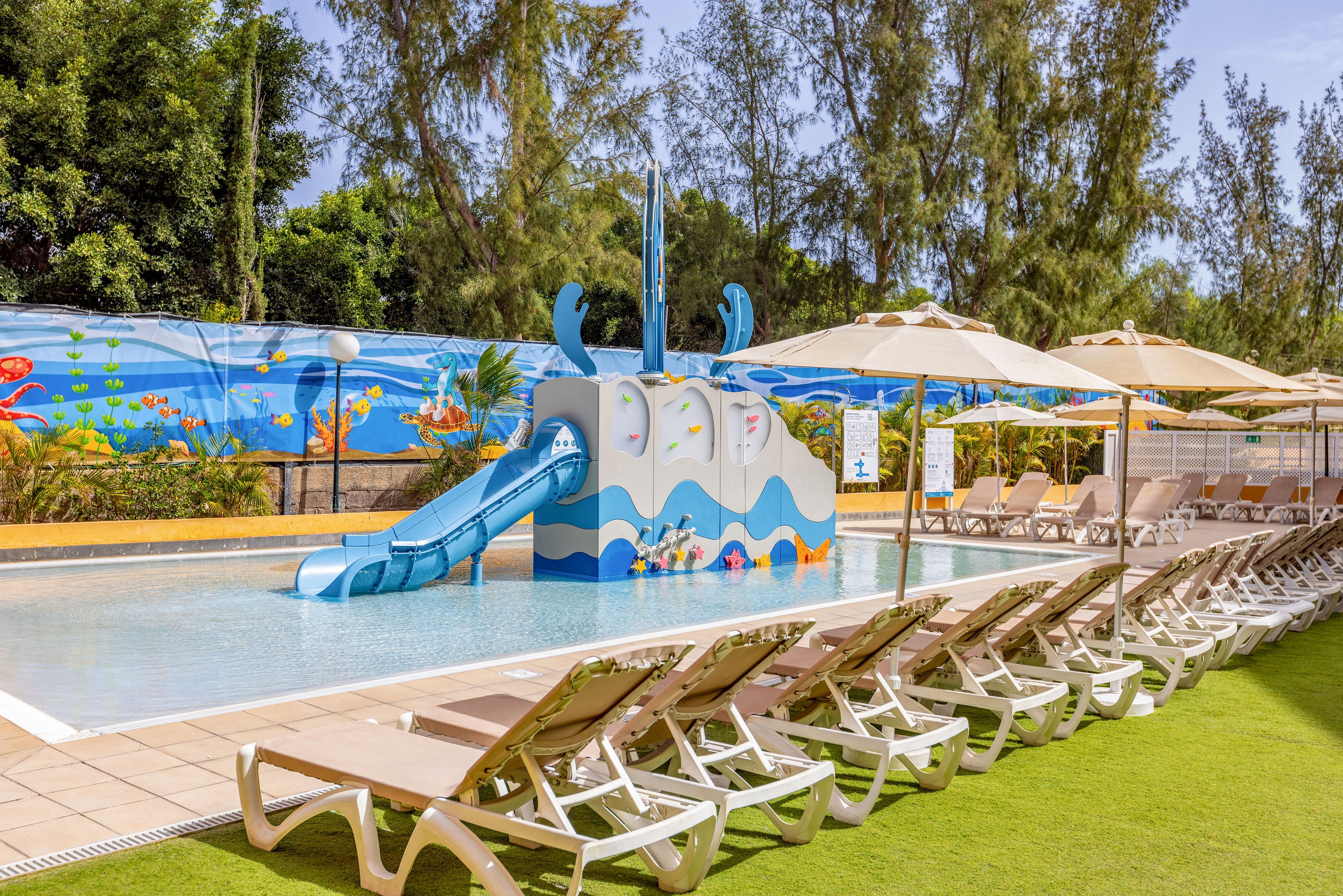 Outdoor children’s swimming pool featuring a playful whale-shaped waterslide is shown on a sunny day. The pool is surrounded by rows of empty lounge chairs and large beige sun umbrellas on a tiled deck and artificial grass. A vibrant underwater-themed mural with fish and sea creatures decorates the wall behind the pool. Tall trees frame the background, creating a relaxed vacation atmosphere.