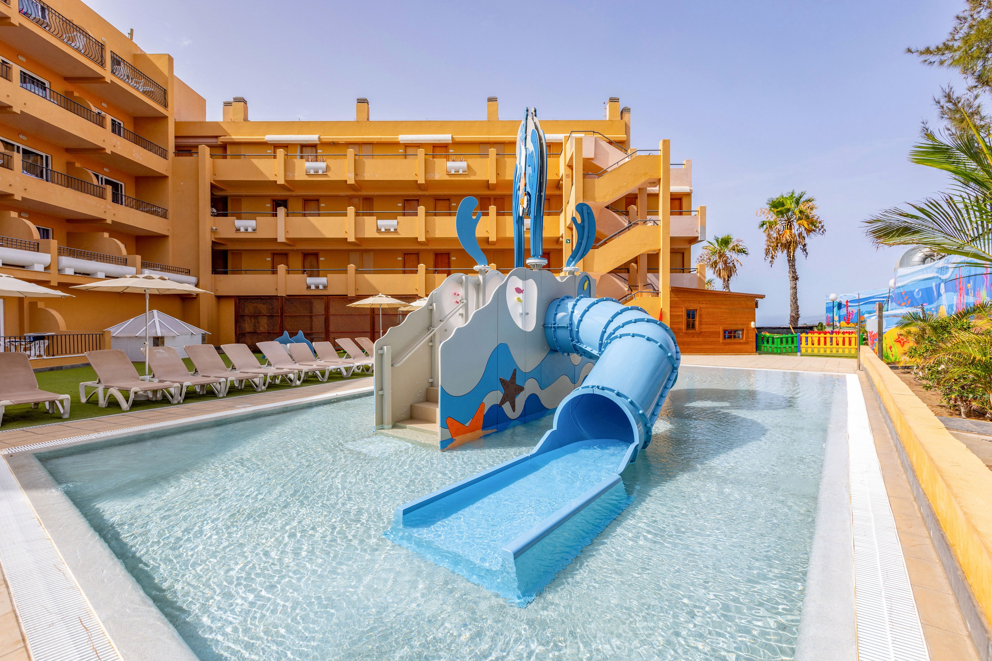 A bright children’s water slide stands in a shallow pool at an outdoor hotel complex. The slide features playful ocean-themed graphics and a blue tube ending in a wide splash ramp. Surrounding the pool are rows of sun loungers and umbrellas, with multi-story yellow buildings in the background. Palm trees and colorful fencing add to the sunny, family-friendly vacation atmosphere.