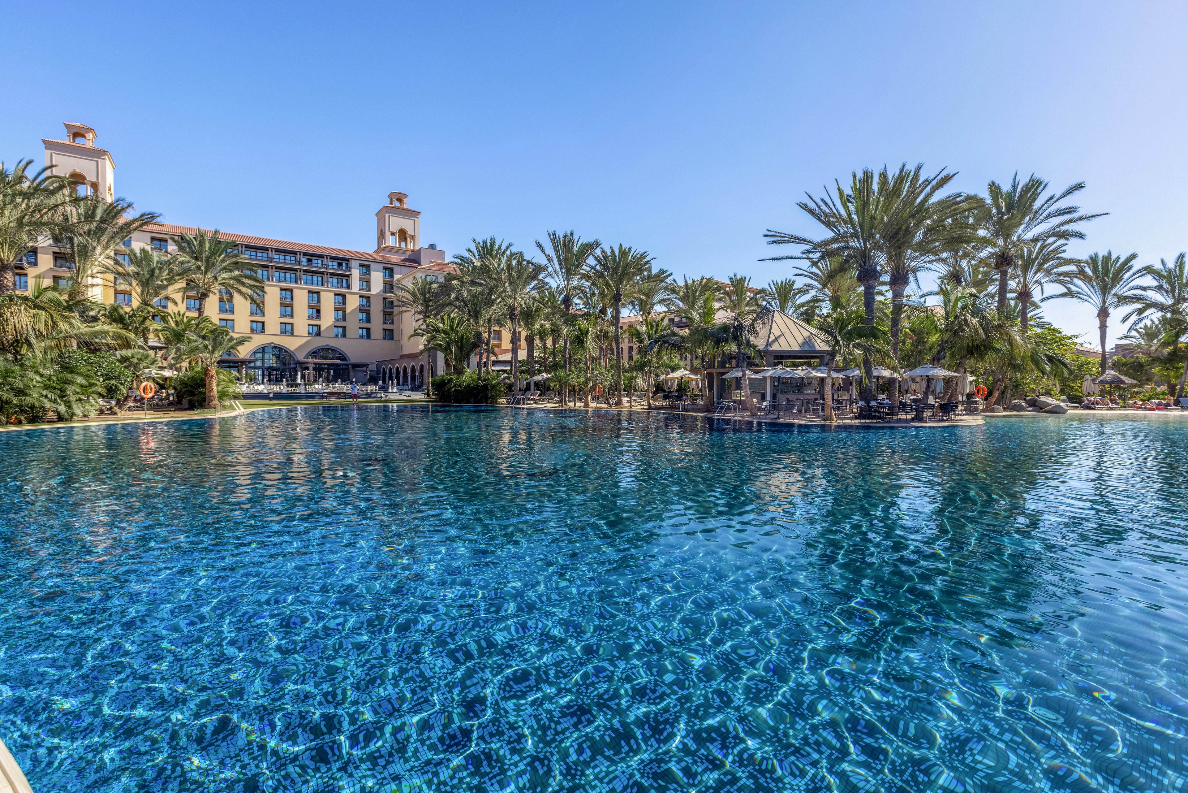 A large resort hotel overlooks an expansive blue swimming pool surrounded by tall palm trees. The clear water reflects the bright sky, creating a vibrant and inviting scene. Guests relax in shaded areas and at a poolside bar nestled among the trees. The architecture of the hotel and the lush landscaping suggest a warm, tropical vacation destination.