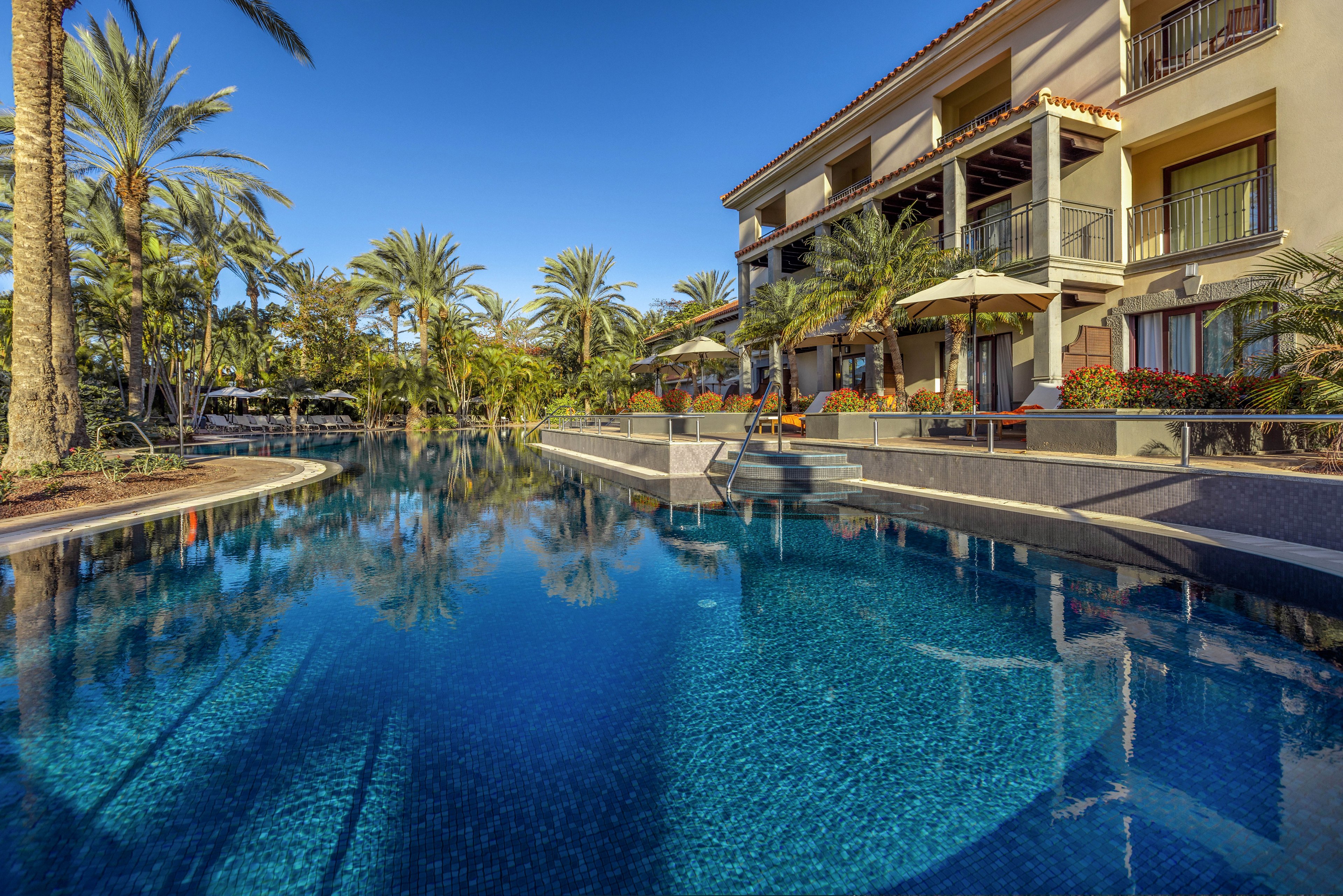 A tranquil resort swimming pool stretches alongside a multi-story hotel building. Tall palm trees and lush greenery surround the water, creating a tropical oasis atmosphere. Sun loungers and umbrellas are arranged on the pool deck, suggesting a relaxing vacation setting. Clear blue skies and reflections in the calm water emphasize the serene, sunny climate.