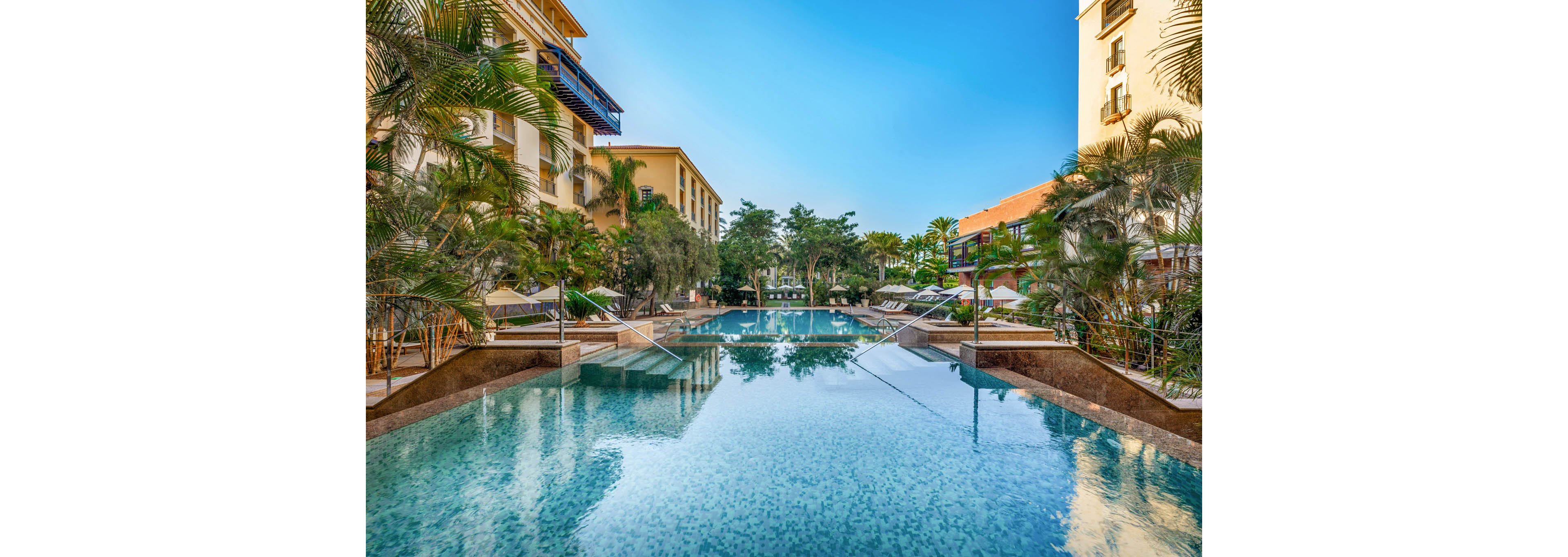 A large, tranquil swimming pool stretches between two resort buildings lined with balconies. Palm trees and lush greenery surround the pool, creating a tropical oasis atmosphere. Sun loungers and umbrellas are arranged neatly along the poolside, suggesting a relaxing vacation setting. The clear blue sky and calm water reflections enhance the serene, sunny mood of the scene.