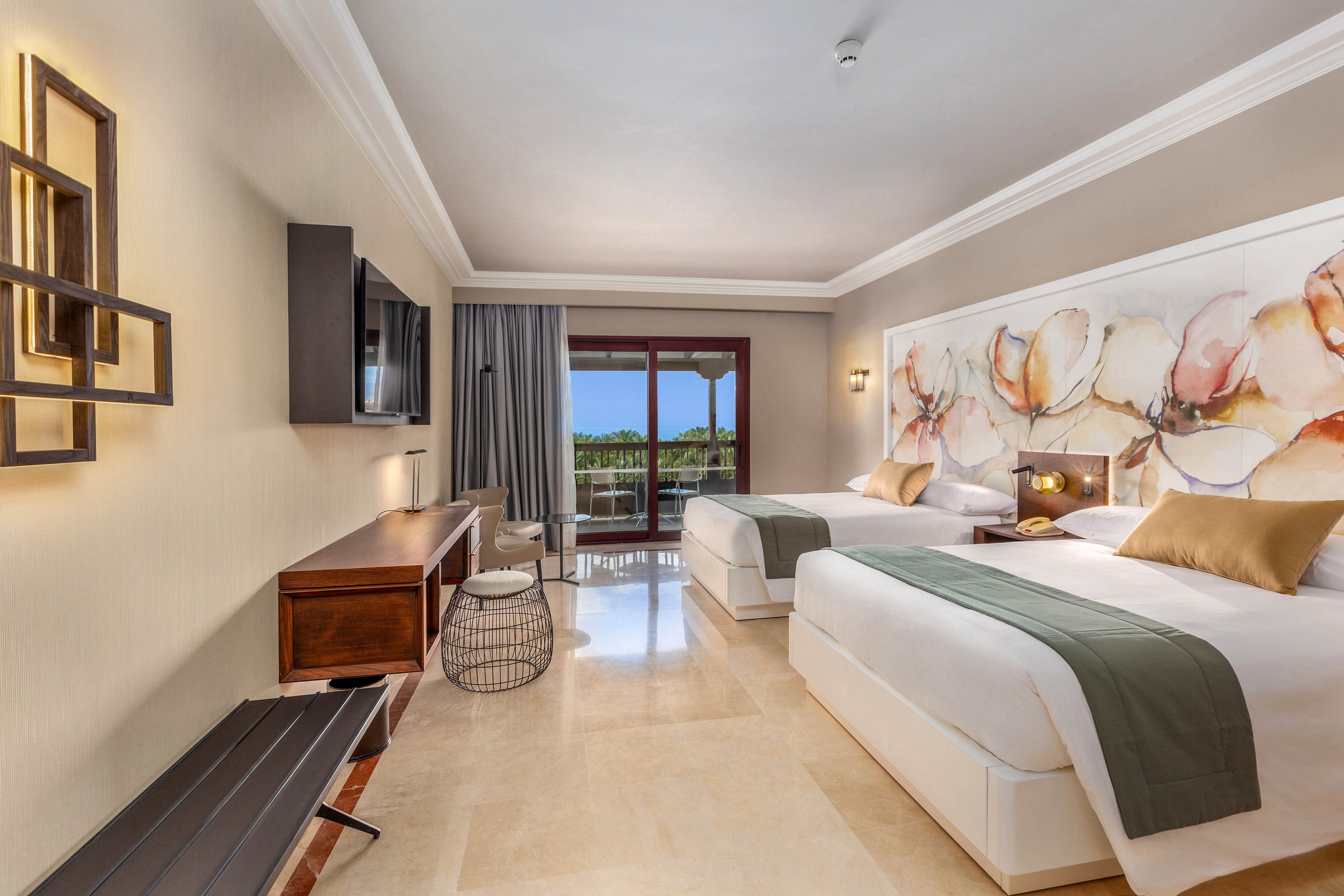 A contemporary hotel room features two neatly made twin beds with white linens, green runners, and beige accent pillows. The space includes a wooden desk with a flat-screen TV, a bench, and a small round stool. Large sliding glass doors open to a balcony with a view of greenery and blue sky. Soft neutral tones, abstract floral wall art, and warm lighting create a calm, inviting atmosphere.