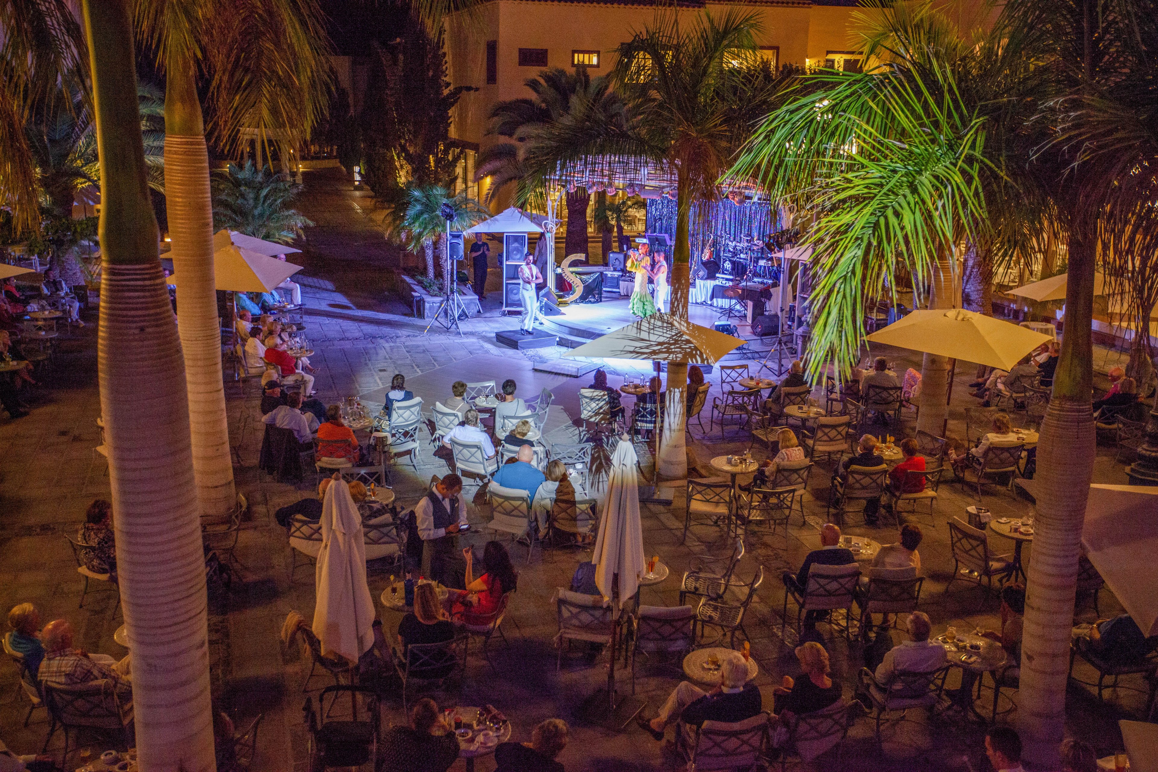An outdoor concert takes place at night in a palm-lined courtyard. Audience members sit at café-style tables under large umbrellas, watching performers on a brightly lit stage. Warm yellow and purple lighting creates a festive atmosphere against the surrounding buildings. The scene suggests a relaxed vacation setting with live music and socializing.