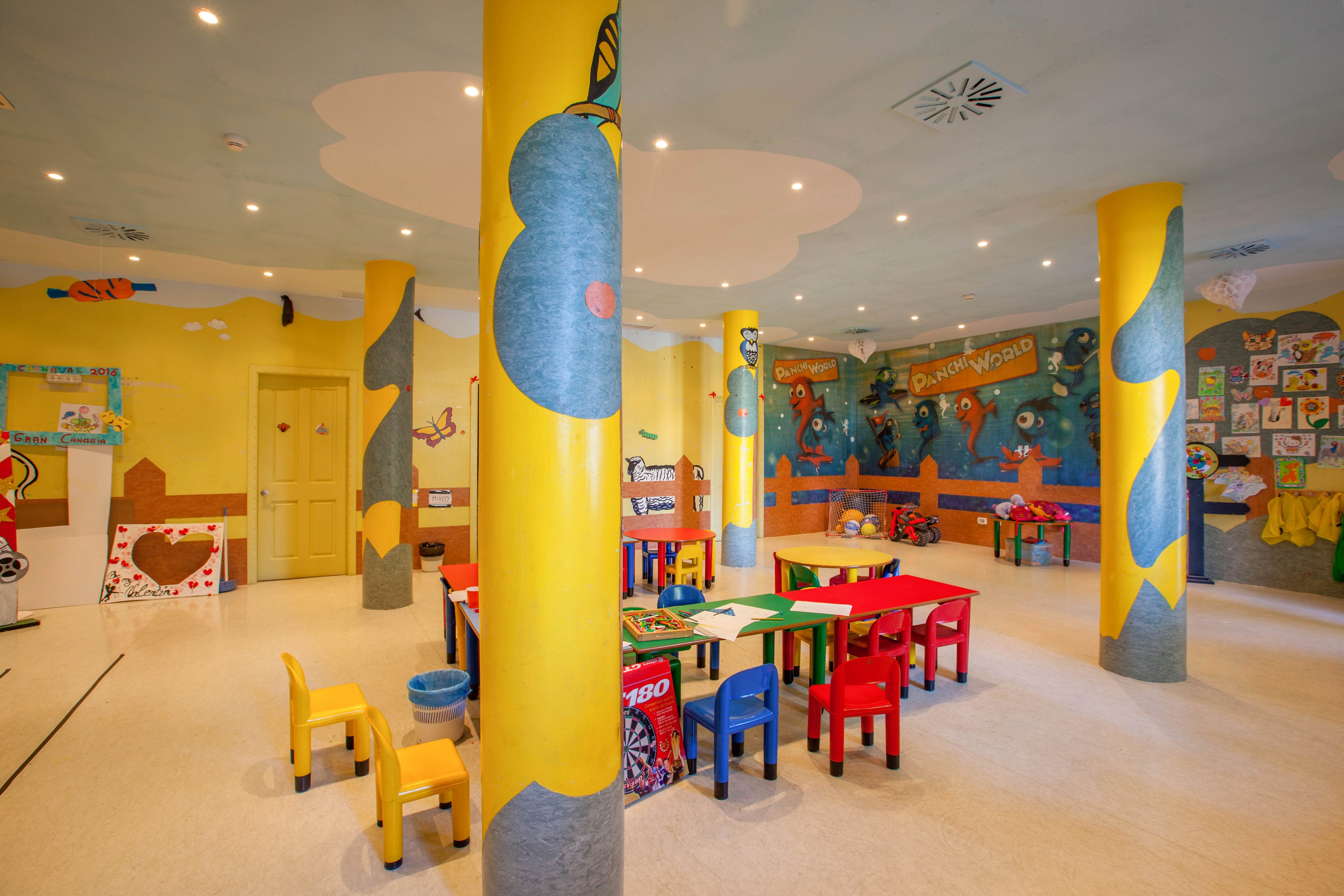 A bright kindergarten classroom features small colorful tables and chairs arranged around the room. Large yellow columns decorated with playful designs stand prominently in the space. The walls are covered with childlike murals, drawings, and crafts, creating a lively and imaginative environment. The overall setting appears clean, organized, and inviting for young children.