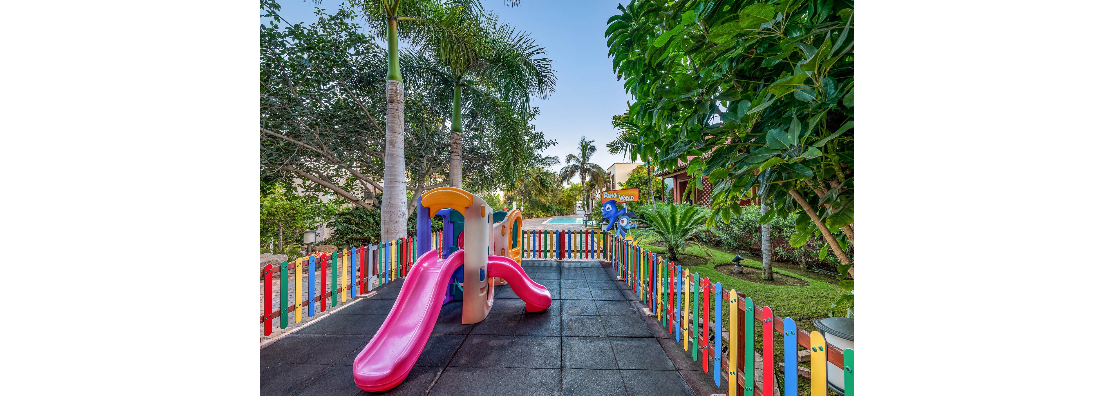 A small playground with a bright pink slide and plastic climbing structure stands inside a fenced area. The fence is made of multicolored wooden slats surrounding a rubber-tiled play surface. Lush green trees and palm trees frame the scene, suggesting a tropical or resort setting. In the background, additional play equipment and landscaped gardens are visible under a clear blue sky.