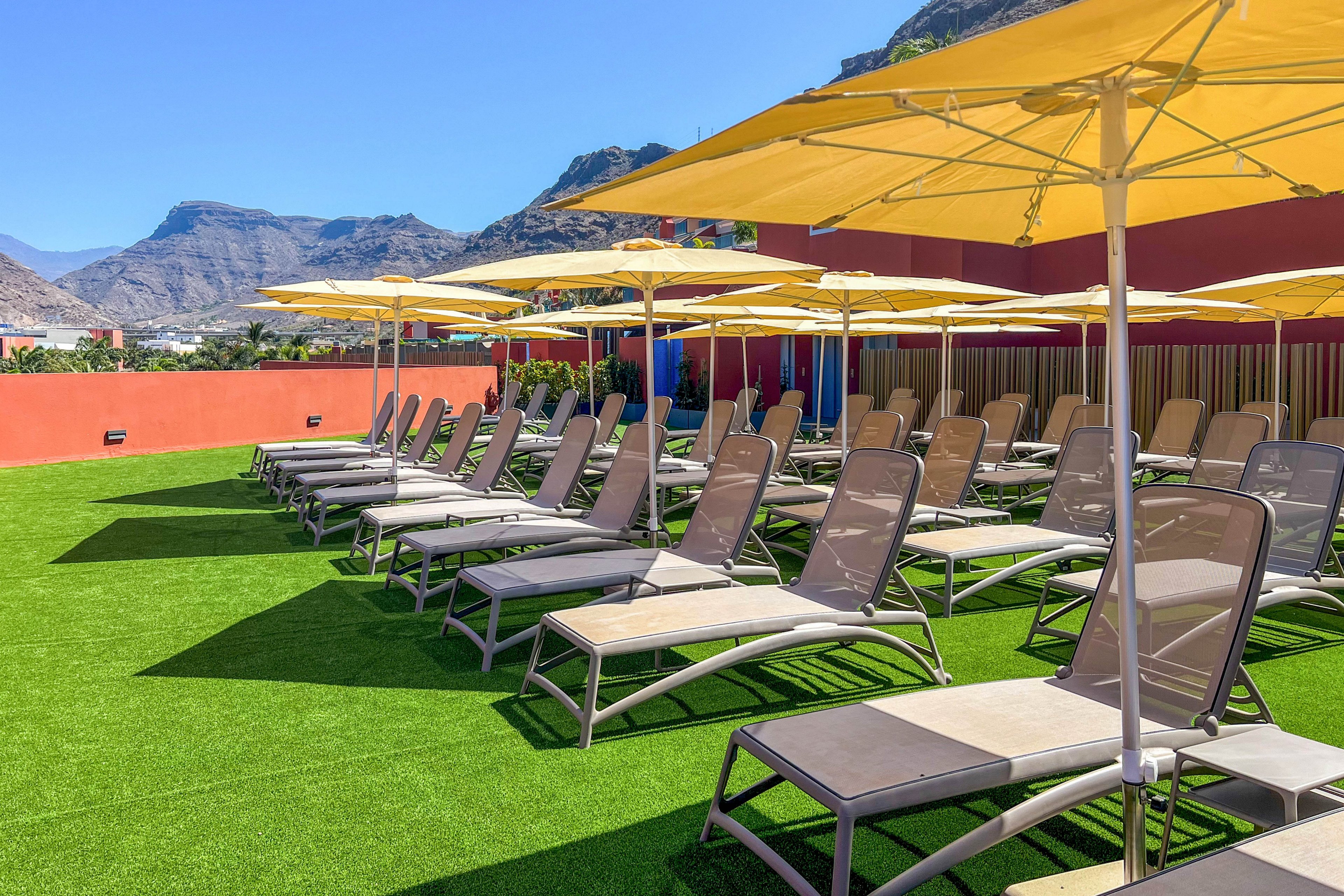 A row of empty sun loungers is arranged neatly on a bright rooftop terrace. Large yellow umbrellas provide shade over the chairs, which sit on a vivid green artificial grass surface. A red wall borders the terrace, with distant mountains and a clear blue sky in the background. The scene suggests a quiet, sunny day at a resort or hotel sun deck.