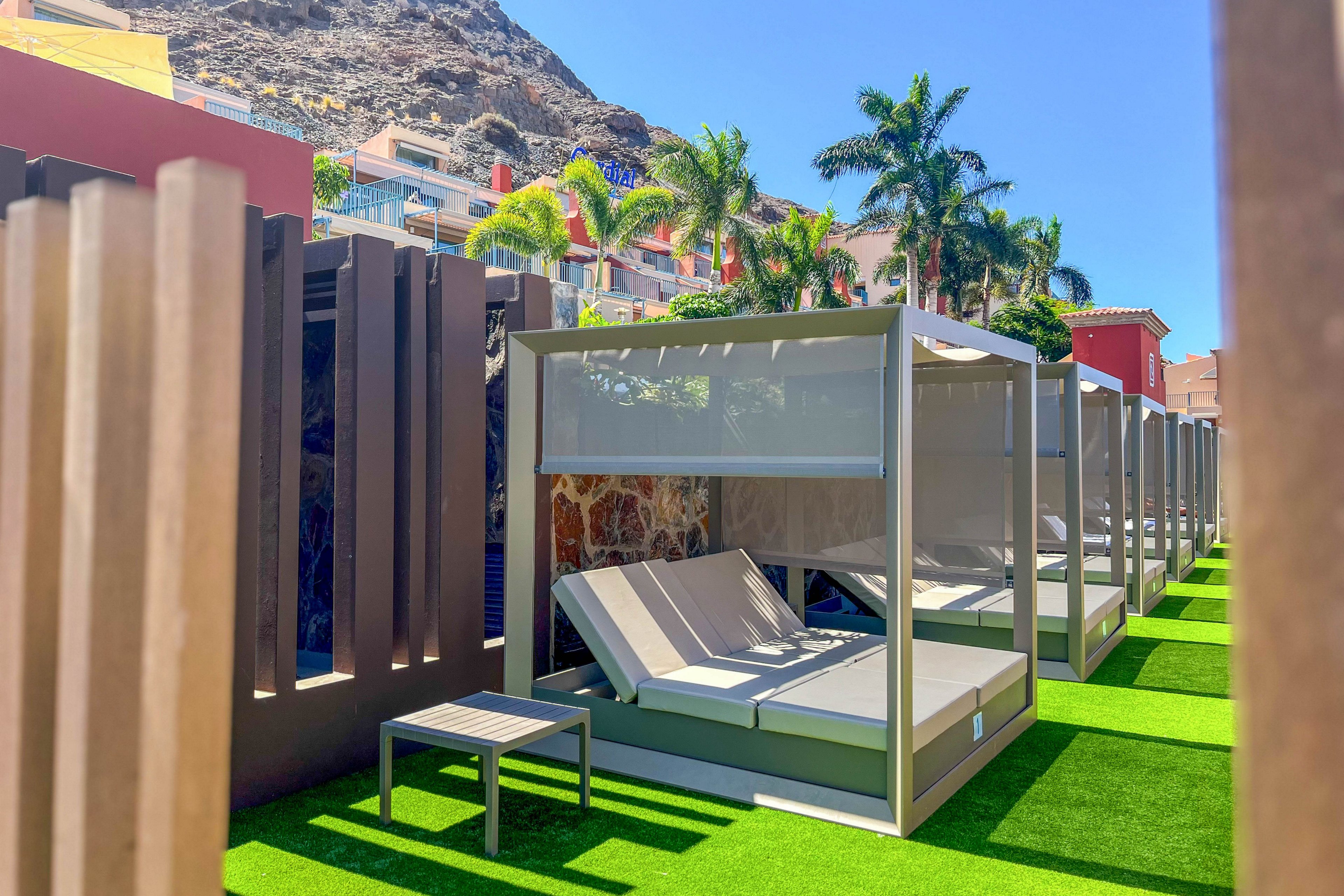 A row of modern cabanas with cushioned sun loungers is arranged on bright artificial grass in an outdoor resort area. The cabanas feature sleek frames and fabric canopies providing shade and privacy. Palm trees and colorful buildings rise in the background beneath a clear blue sky. The scene suggests a sunny, relaxing vacation setting with contemporary design elements.