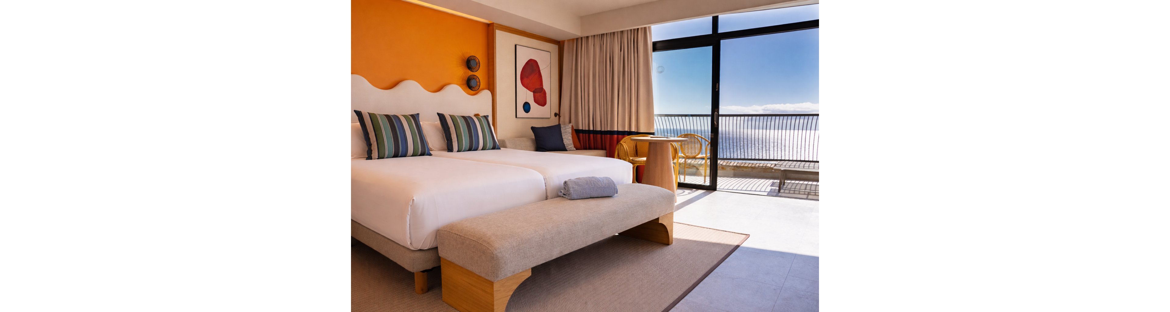 A contemporary hotel room features a large double bed with striped pillows and a cushioned bench at the foot. Warm orange and neutral walls are accented by modern artwork and decorative wall plates. A sliding glass door opens to a balcony with a view of the ocean and clear sky. Natural light fills the room, highlighting the cozy seating area with a small table and chairs near the window.