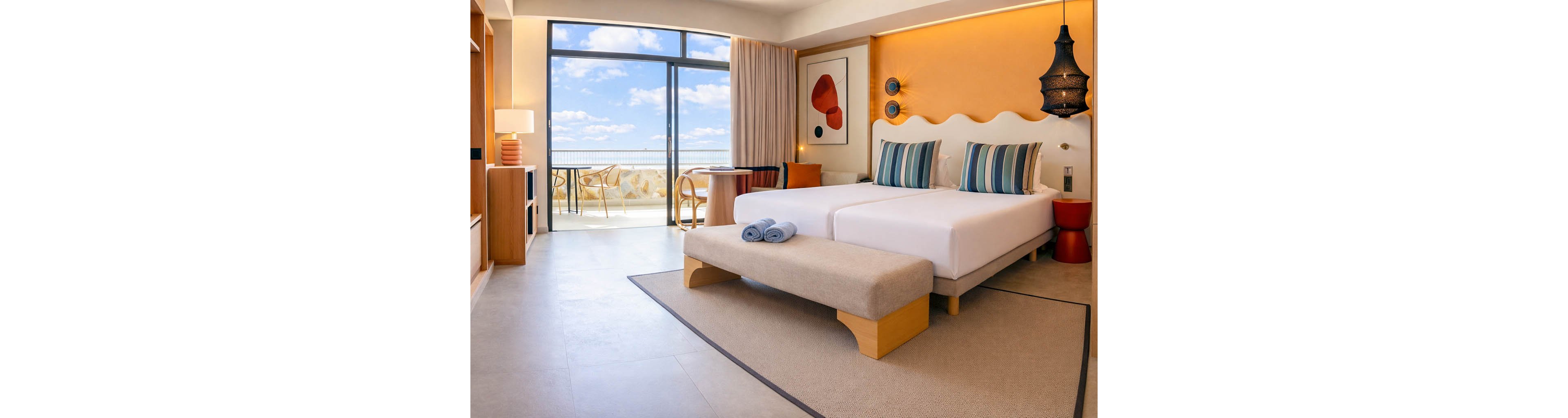 A contemporary hotel room features two neatly made twin beds with striped accent pillows and a cushioned bench at the foot. Warm orange and neutral tones dominate the decor, complemented by abstract wall art and a sculptural black pendant light. Large glass doors open to a balcony with outdoor seating and a scenic view of the sky and distant landscape. Soft natural light fills the space, creating a calm and inviting atmosphere.