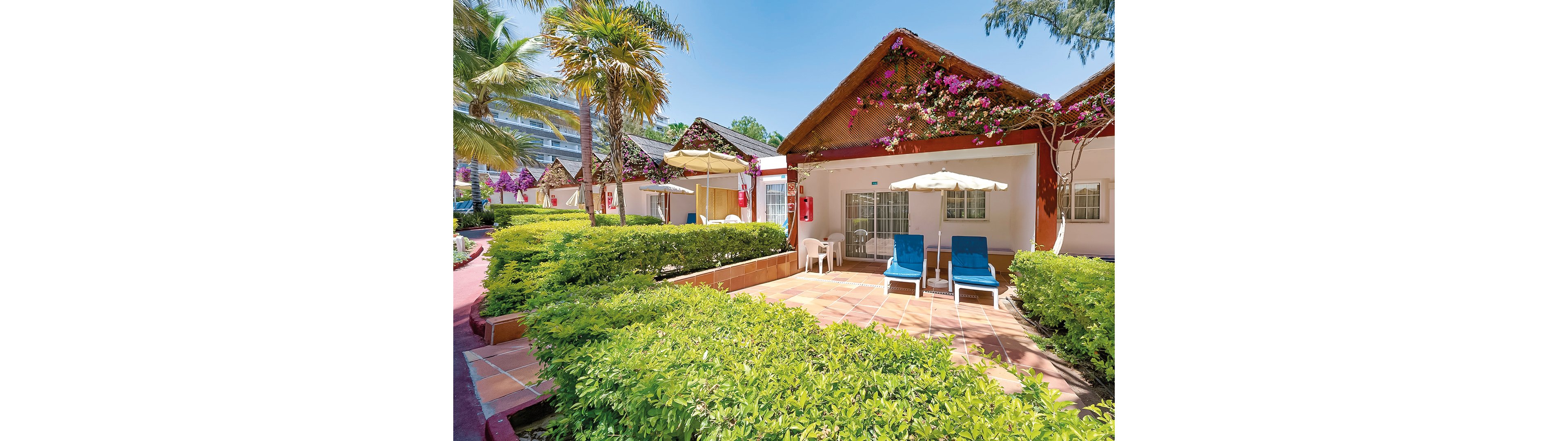 A serene view of tropical resort villas surrounded by vibrant greenery and blooming flowers. The setting features tiled patios with lounge chairs and umbrellas, ideal for relaxation. Palm trees and bright blue skies enhance the tranquil atmosphere, creating a picturesque vacation spot.