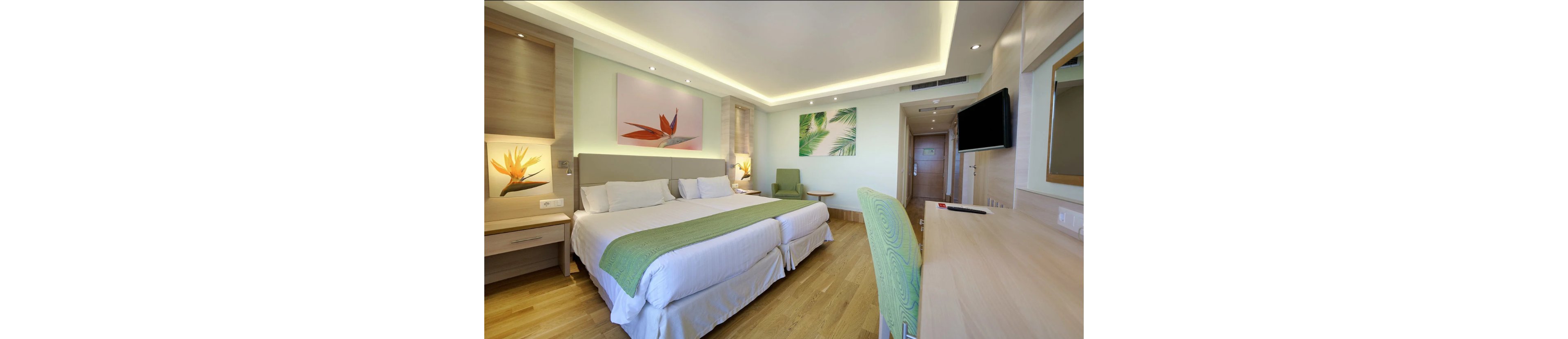 Bright contemporary hotel room featuring two neatly made twin beds with green runners. The space includes light wood furniture, a desk with chair, and a wall-mounted flat-screen television. Soft indirect ceiling lighting and pastel green walls create a calm, inviting atmosphere. Decorative artwork with tropical leaf and flower motifs adds color above the beds and seating area.