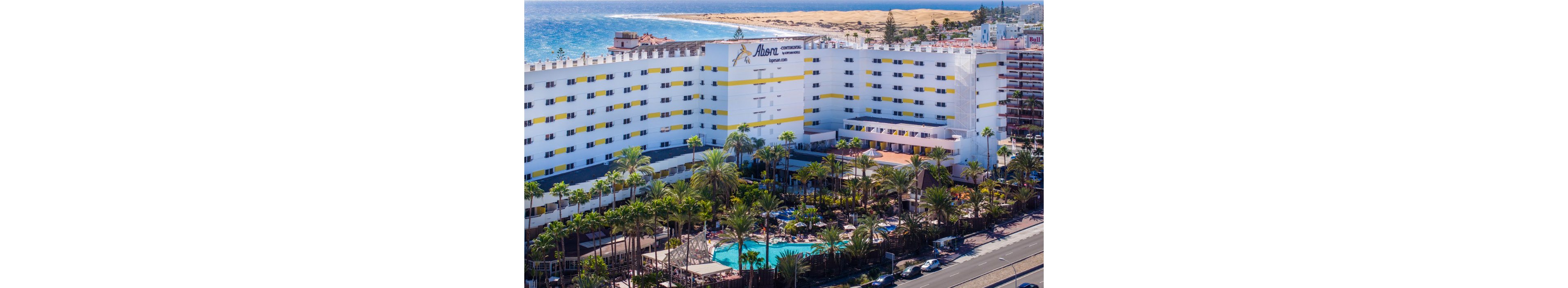 A large multi-story resort hotel stands near the coastline, with the ocean and sandy dunes visible in the background. The foreground features lush palm trees surrounding an outdoor swimming pool area filled with sun loungers and umbrellas. A main road runs in front of the property, with additional buildings and accommodations nearby. The scene is captured on a clear, sunny day, emphasizing a vibrant vacation atmosphere.