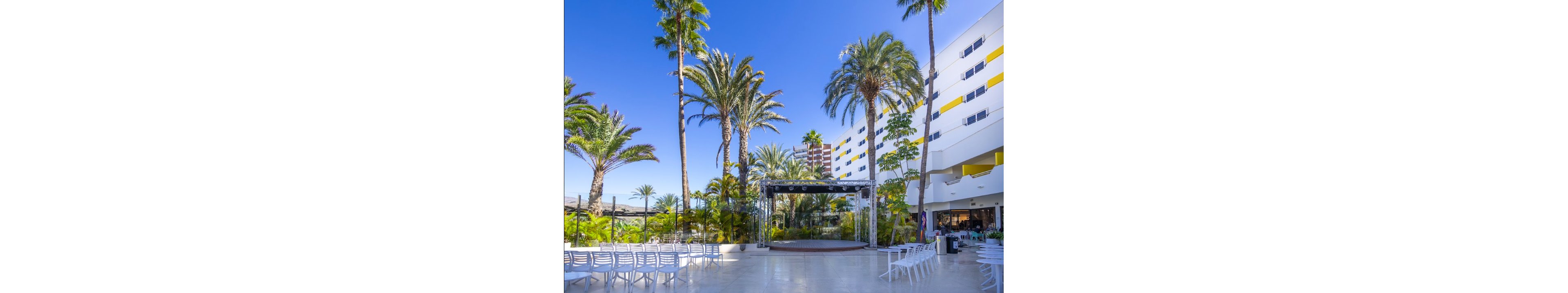 An open-air event area is set up with rows of white chairs facing a small stage structure. Tall palm trees and lush greenery surround the space, creating a tropical atmosphere. A modern multi-story hotel building with white and yellow accents borders the courtyard. The bright blue sky and strong sunlight give the scene a warm, inviting feel, suitable for gatherings or performances.
