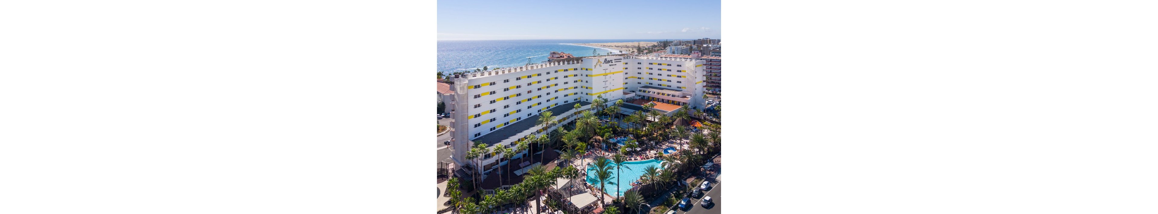 A large multi-story resort hotel stands near a sandy coastline overlooking the ocean. The building features white walls with yellow accents and a central tower section, surrounded by lush palm trees. In the foreground, a bright blue outdoor swimming pool is filled with guests relaxing on loungers and under umbrellas. The surrounding area includes nearby streets, parked cars, and distant beachfront buildings under a clear sky.