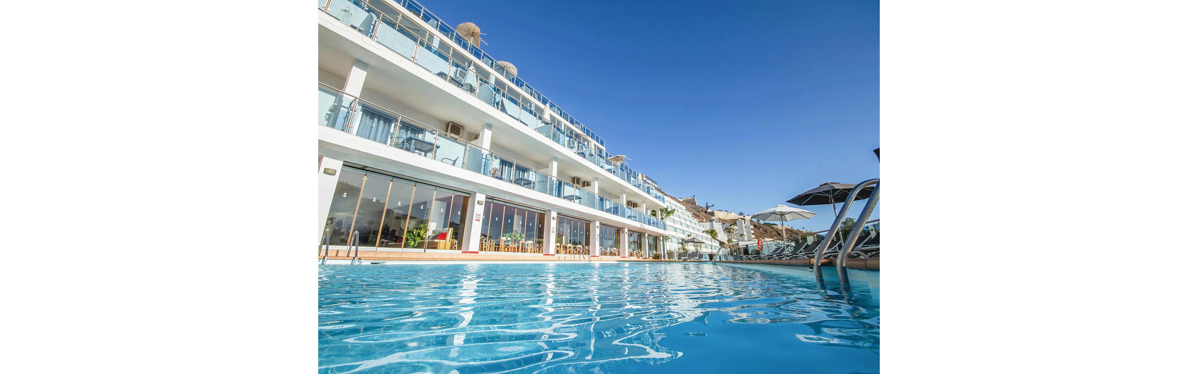 A bright blue swimming pool stretches across the foreground of a modern multi-story hotel. The building features glass balconies and large windows overlooking the water. Sun loungers and umbrellas line the poolside area, suggesting a relaxing vacation setting. Clear skies and strong sunlight create a warm, inviting atmosphere at this coastal resort.