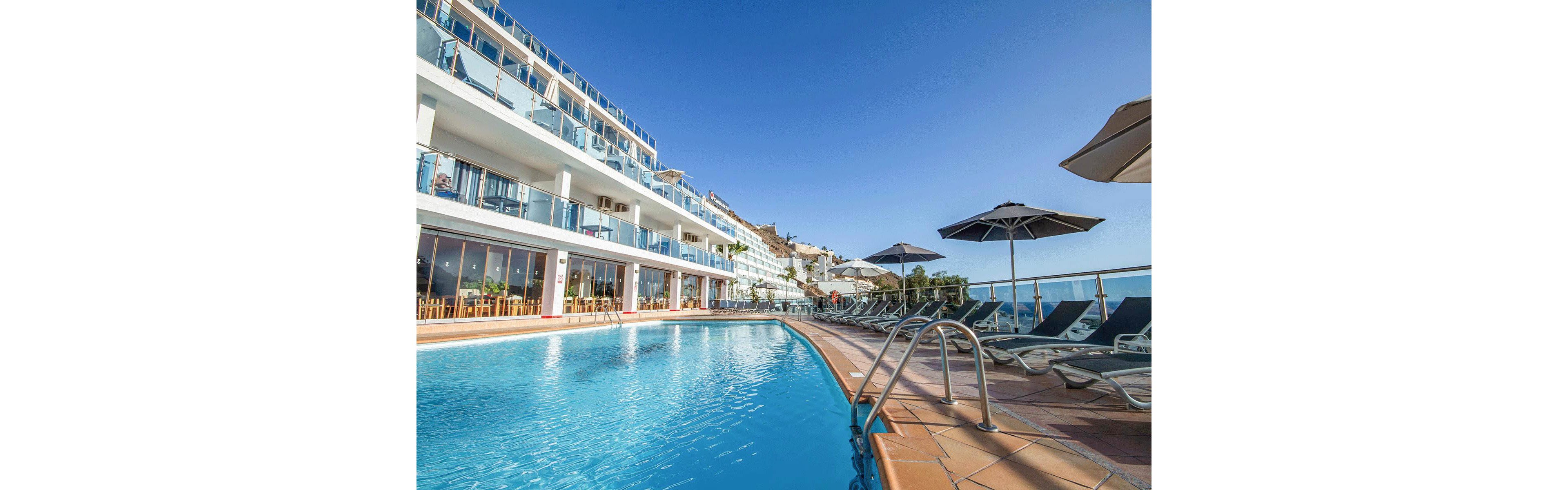 A contemporary multi-story hotel overlooks a large outdoor swimming pool under a clear blue sky. Sun loungers and umbrellas line the poolside terrace, facing toward the ocean view. Glass balconies and wide windows reflect the bright daylight, emphasizing the resort’s modern architecture. The scene suggests a relaxing vacation atmosphere in a coastal destination.